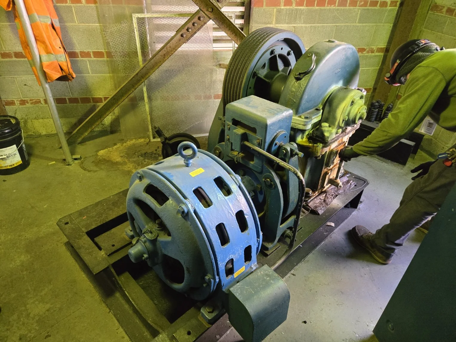 A worker inspecting or repairing industrial machinery, which consists of large interconnected components and gears, in a workshop with brick walls and various tools and materials around.