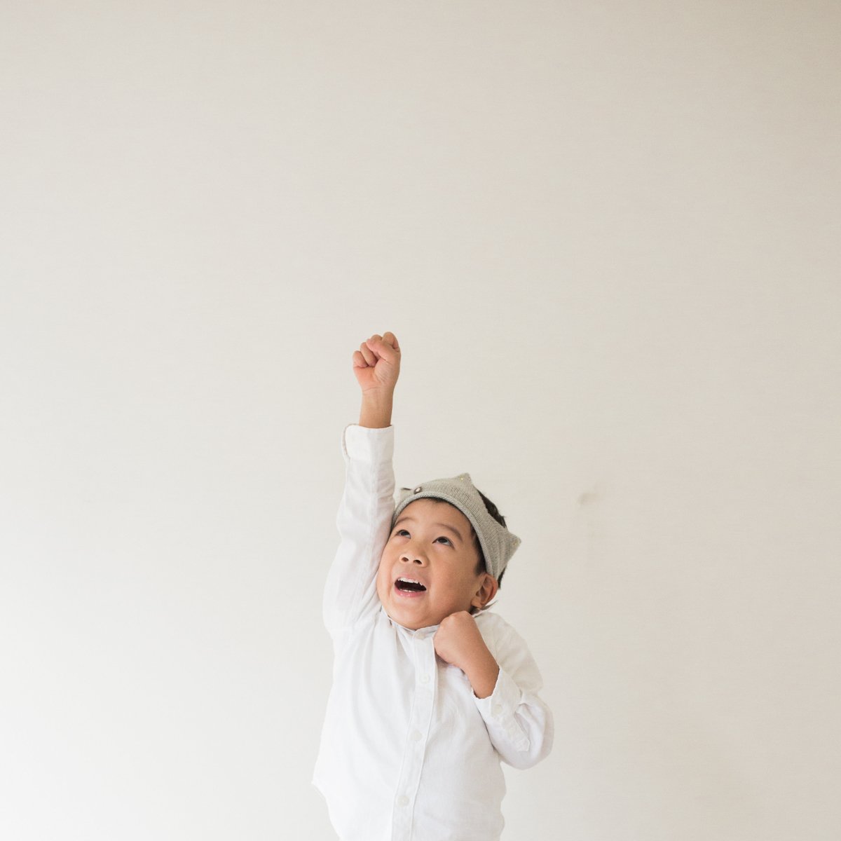 A young boy in a white shirt wearing a gray paper crown raising his arm in excitement against a plain light background.