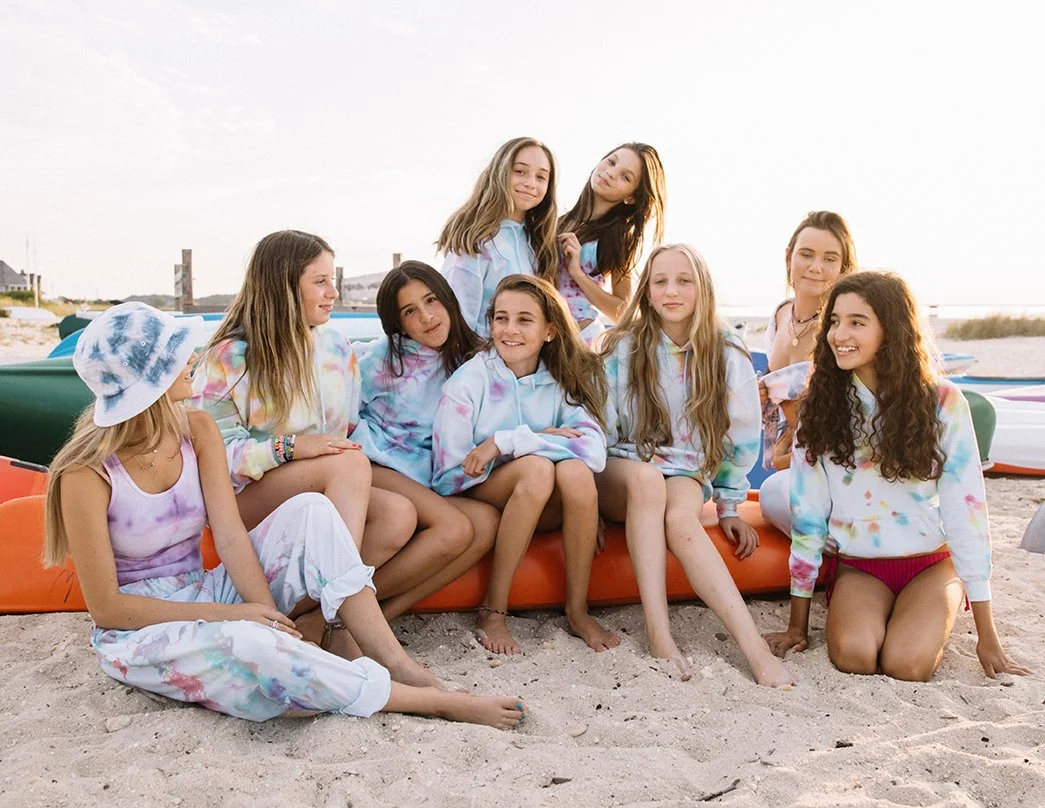 Group of girls sitting and standing on a sandy beach with colorful kayaks in the background, some wearing tie-dye hoodies and others in swimsuits.