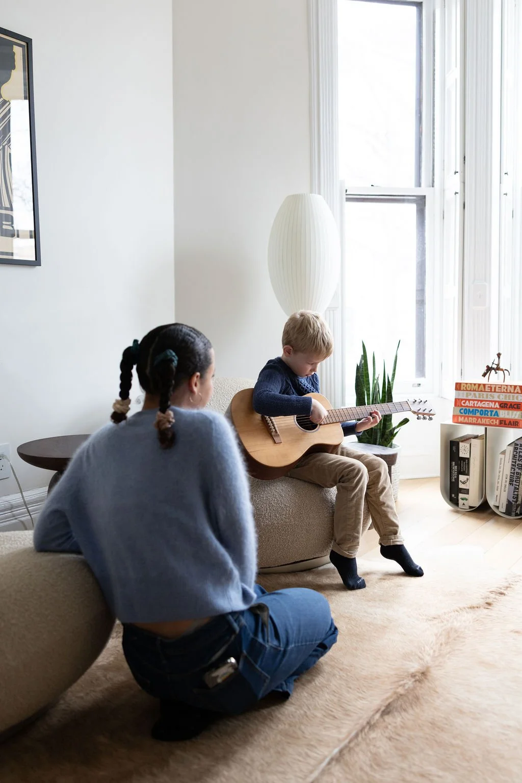A young boy playing an acoustic guitar while sitting on a beige ottoman in a bright living room. A girl with braided hair, wearing a gray sweater, is crouching and observing him. The room has large windows, a white lamp, a potted plant, and stacked b