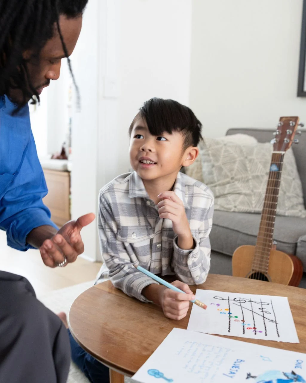 A young boy with dark hair sitting at a round wooden table with a pencil in his hand, smiling and talking to an adult man with dreadlocks, in a living room with a guitar against a sofa in the background.