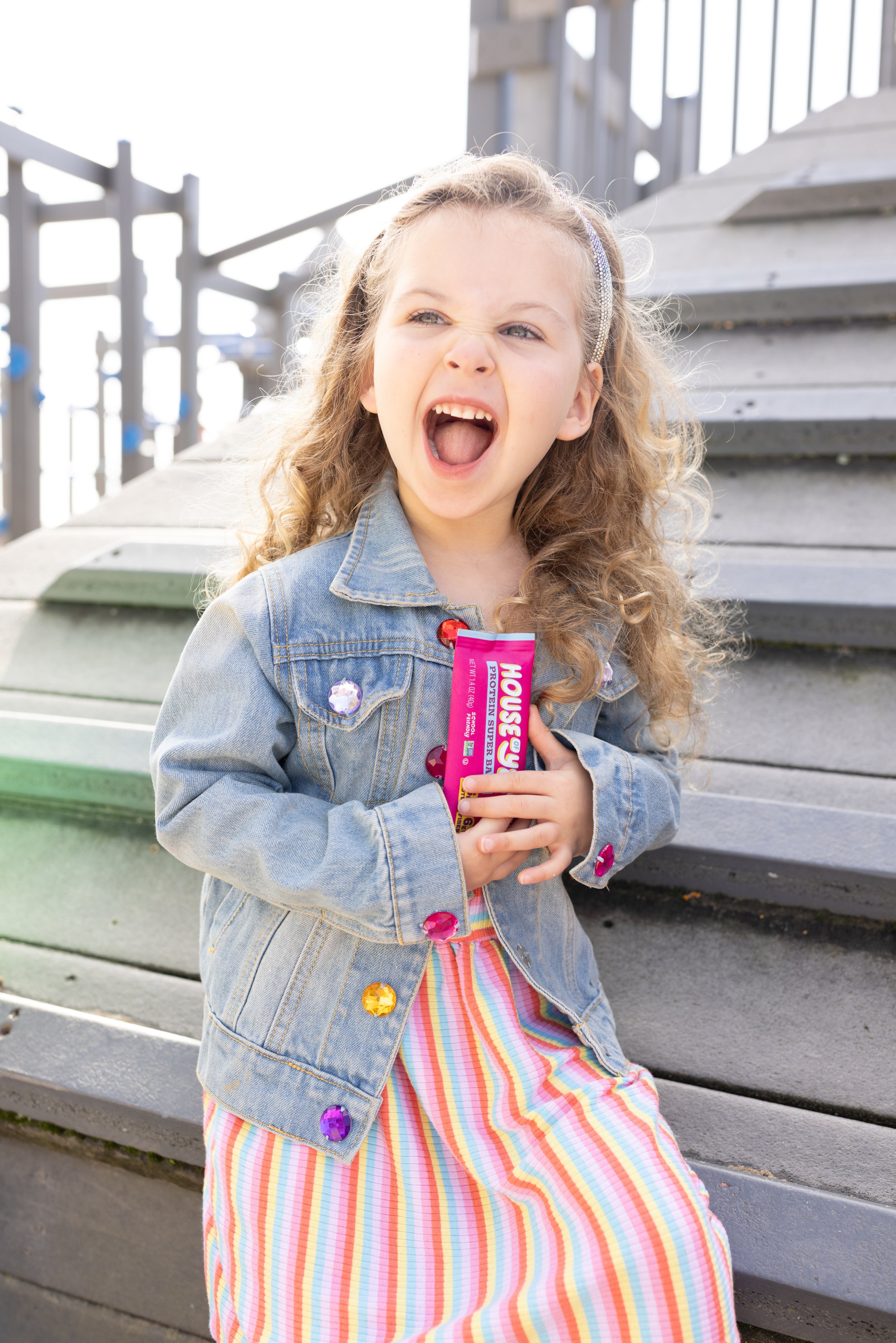 A young girl with light hair and a denim jacket, sitting on steps outdoors, holding a pink pack of Gushers candy, with an expressive, playful face.
