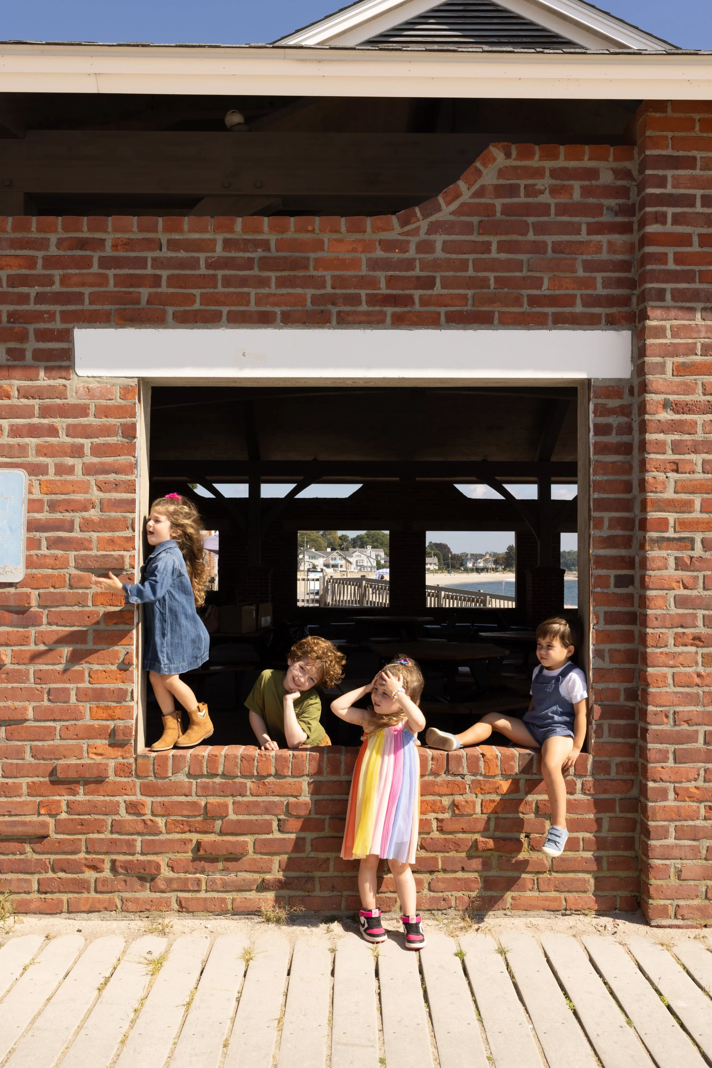 Children playing and sitting on a brick wall structure near water with houses in the background. One girl is climbing out of the wall, others are sitting or standing, some with playful poses, in a casual outdoor setting.