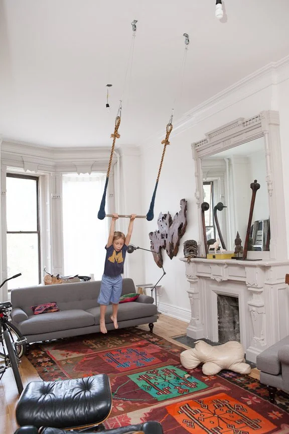 Child swinging on a homemade trapeze in a living room, with a gray sofa, colorful rug, and decorative items on the mantle.