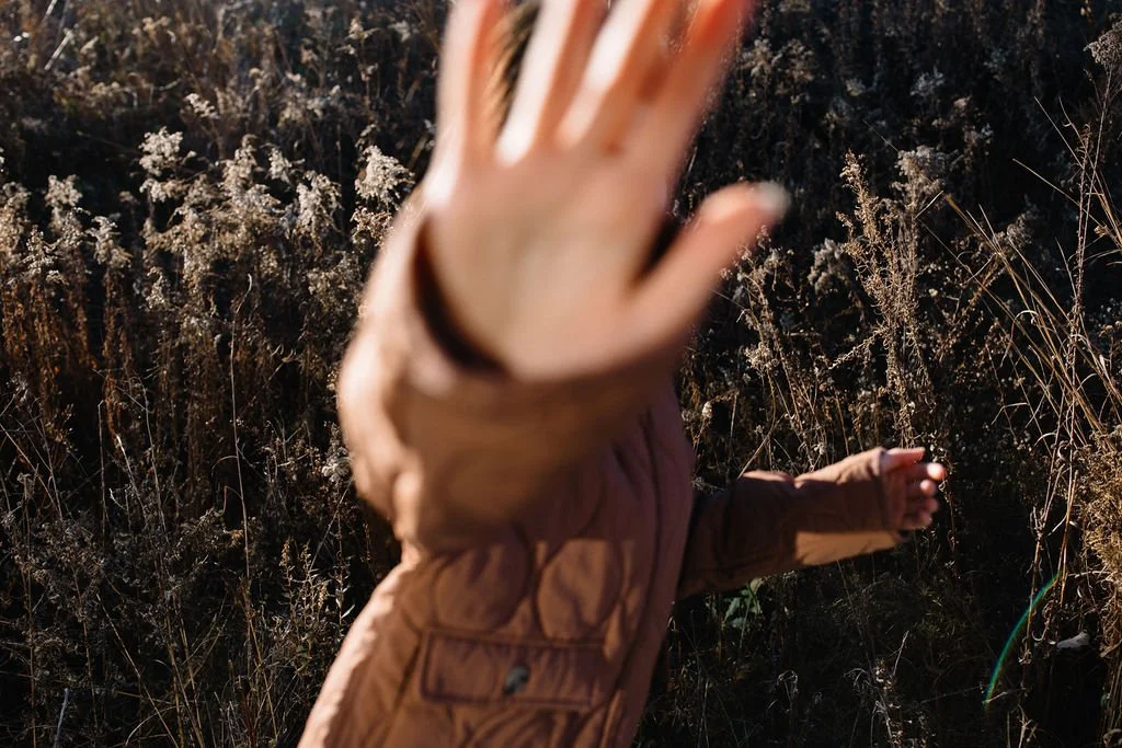 Child in a brown jacket reaching out with hand in front of the camera, standing in a field with tall dry grass.