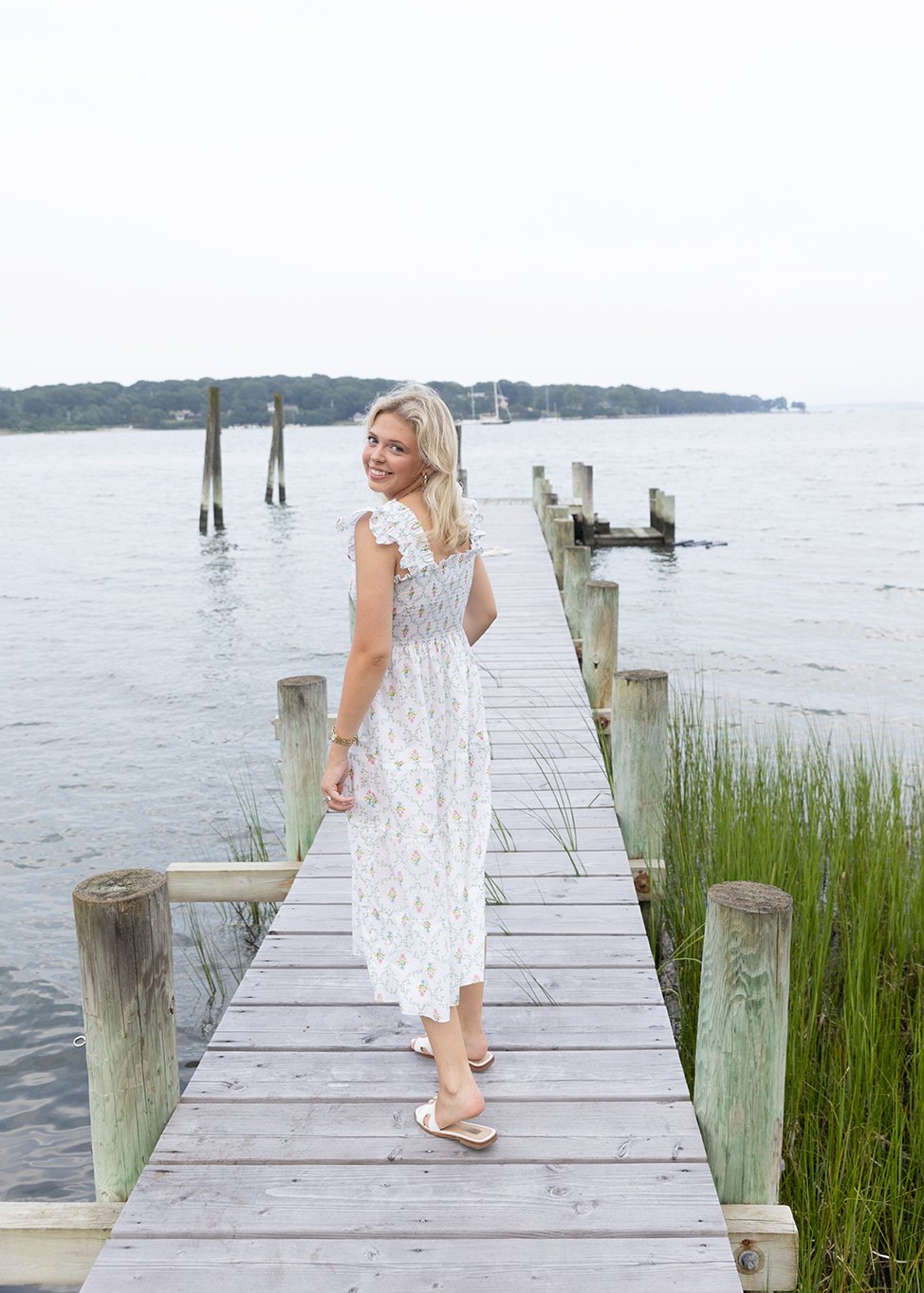 A woman in a white floral dress standing on a wooden dock by the water, smiling and looking back, with a boat and a distant shoreline in the background.