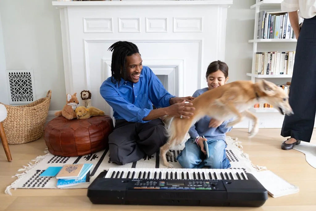 A joyful man, a young girl, and a dog playing together on a rug in a living room. The man is sitting on the floor, smiling and reaching towards the dog, which is mid-leap. The young girl is sitting next to him, holding a leash. There are books on a b