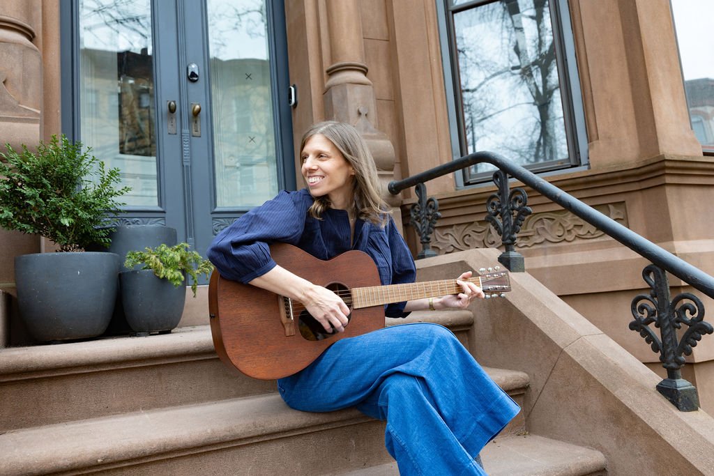 A woman with long hair sitting on outdoor steps of a brownstone, playing an acoustic guitar, with potted plants beside her.