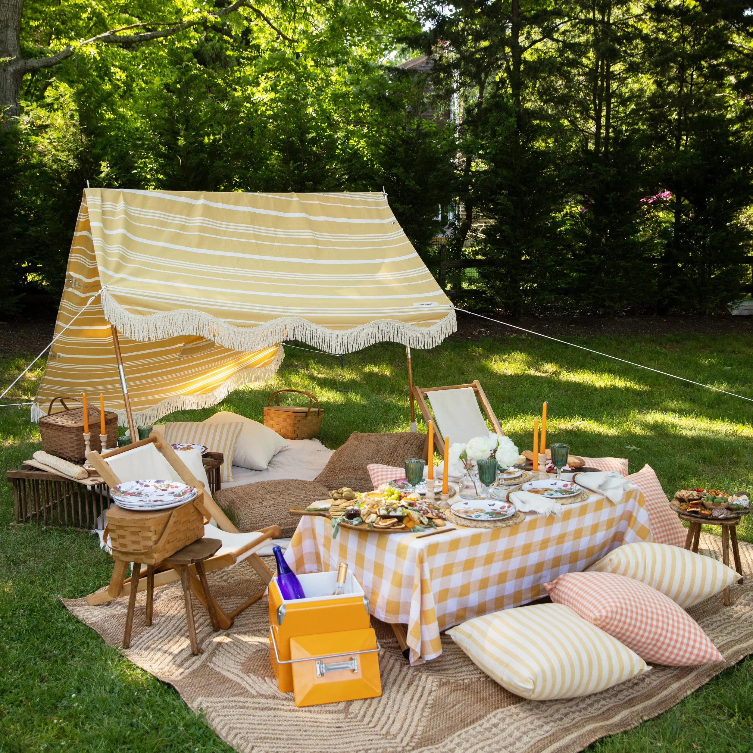 Outdoor backyard picnic setup with a table covered with a yellow checkered tablecloth, outdoor cushions, and a variety of food including cheese and fruits, surrounded by chairs, baskets, and decorative candles on a grassy area under trees.