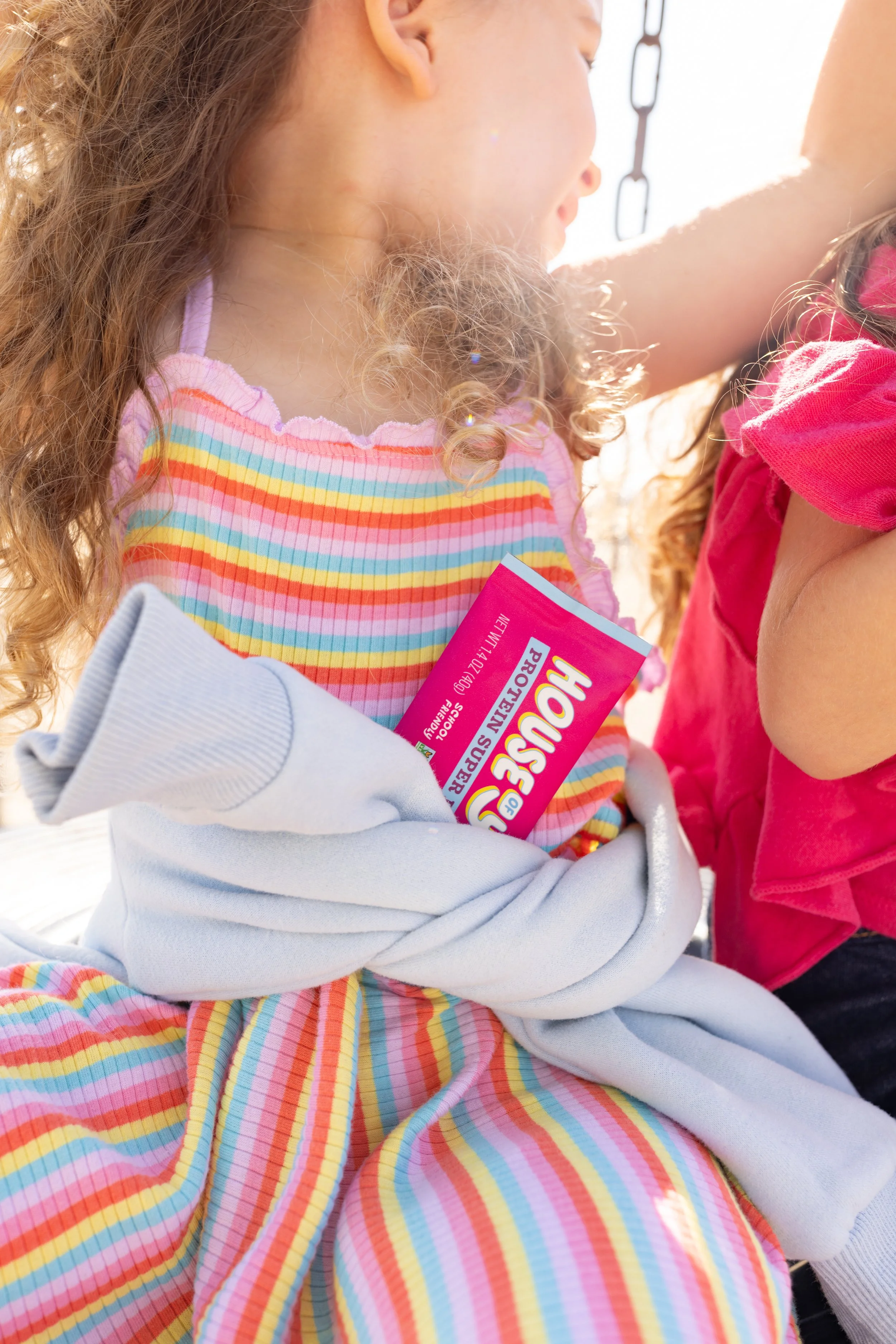 Close-up of a young girl with curly hair wearing a colorful striped shirt holding a pink Hershey's chocolate bar outside in sunlight.
