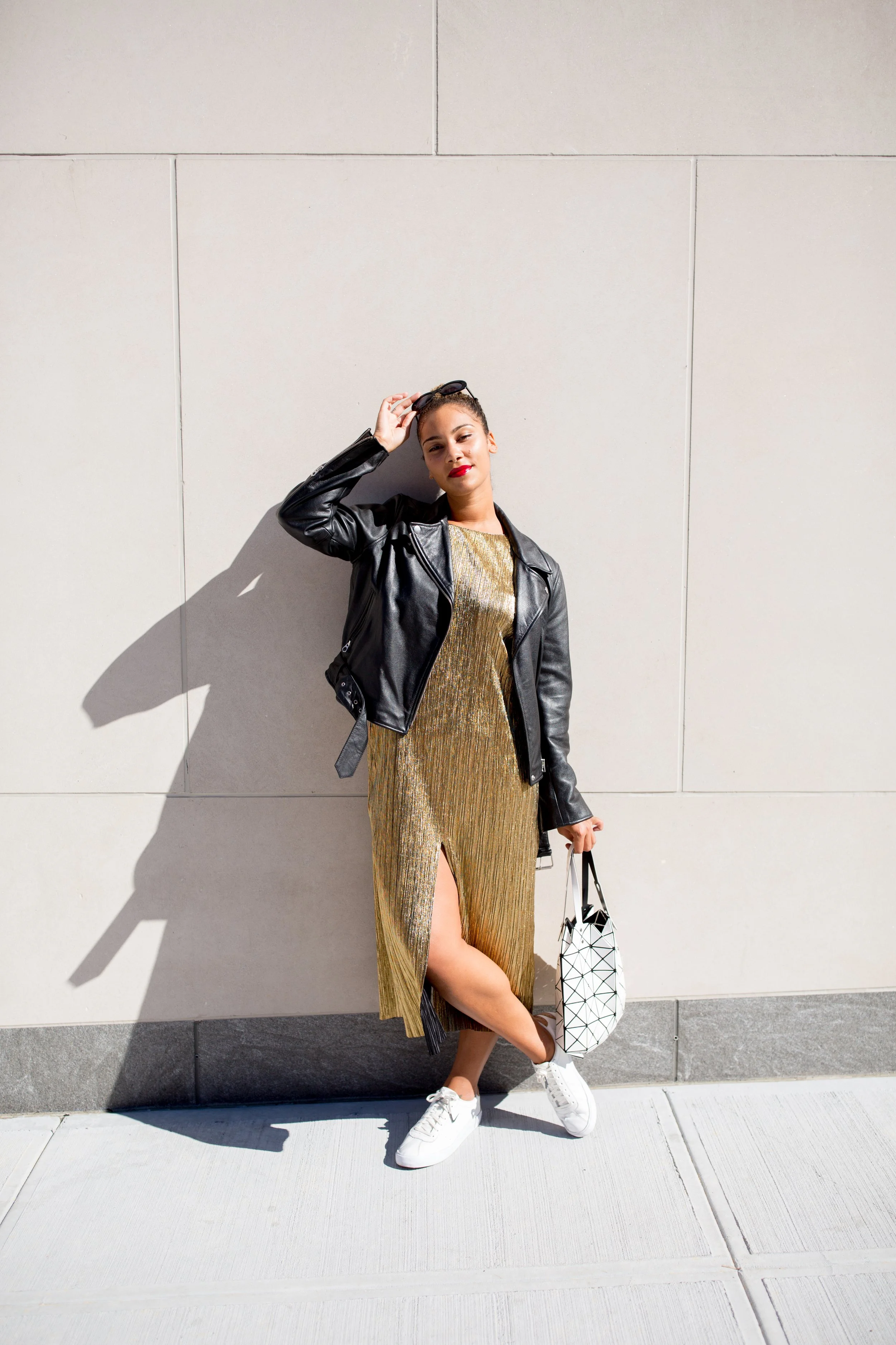 Woman in a gold dress with a slit, white sneakers, black leather jacket, holding sunglasses and a geometric patterned white bag, standing against a plain white wall with strong shadow.