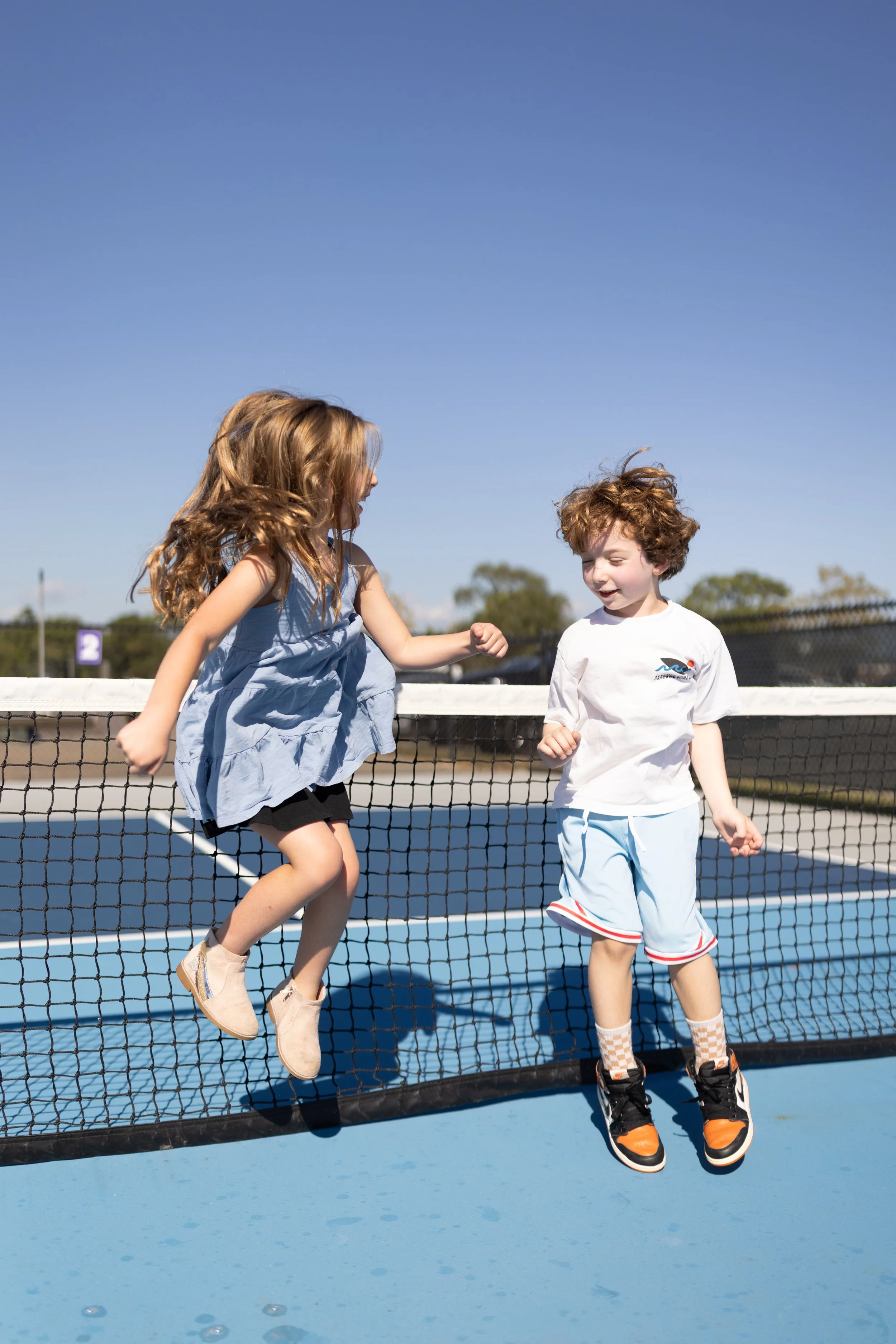 Two children playing on a tennis court, with the girl jumping and the boy standing, both smiling.