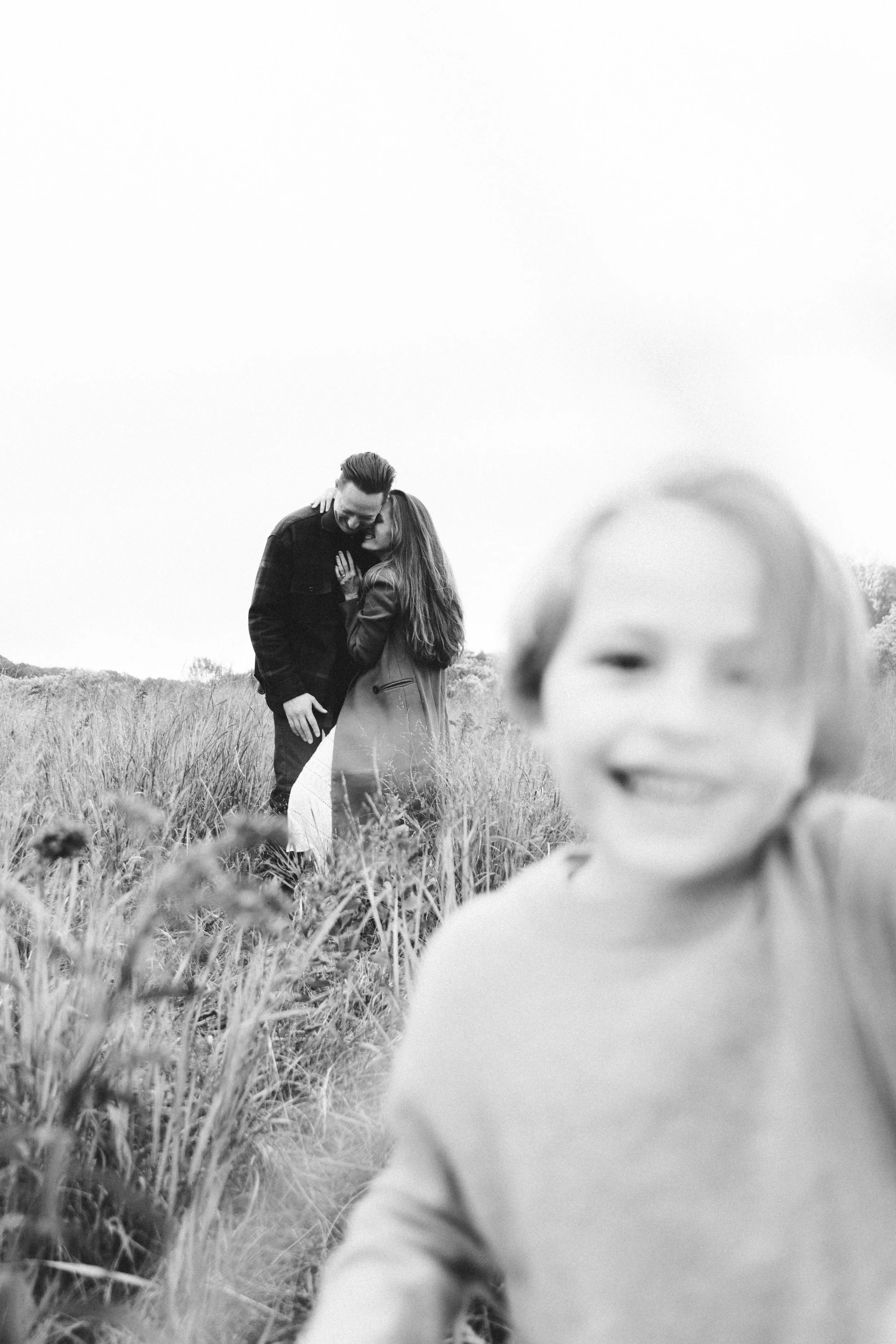 A black and white photo of a man and woman embracing in a field, with a blurred child in the foreground.