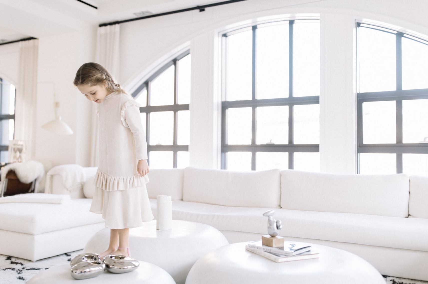 A young girl with long hair in a braid, wearing a cream-colored dress, stands on a white ottoman in a modern, bright living room. The room has large arched windows, white furniture, and decorative objects.