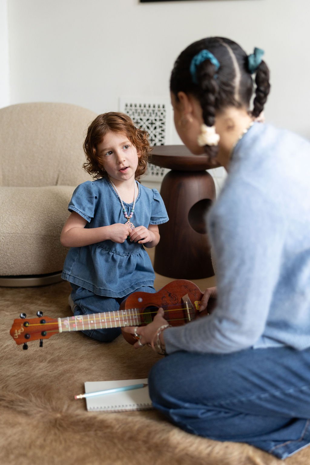 A young girl with curly red hair, wearing a blue dress and a necklace, listens as an older girl with braids and hair accessories plays a small guitar.