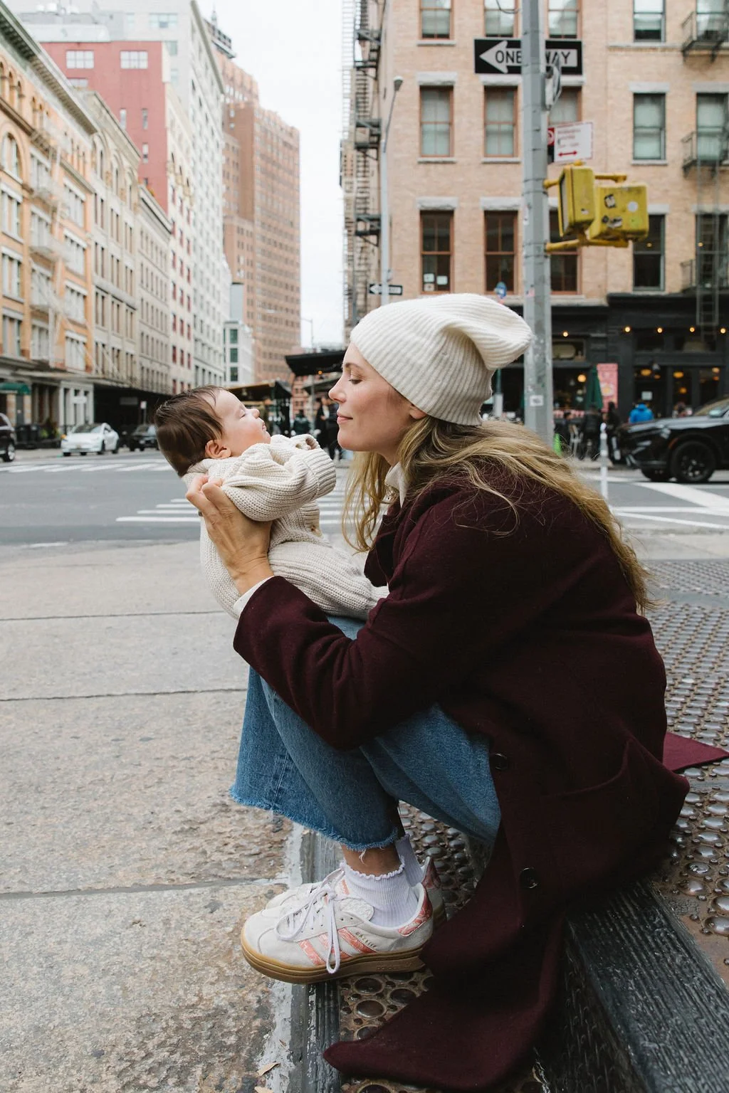 A woman in a beige beanie and maroon coat holding a young child in a cream sweater on a city sidewalk.