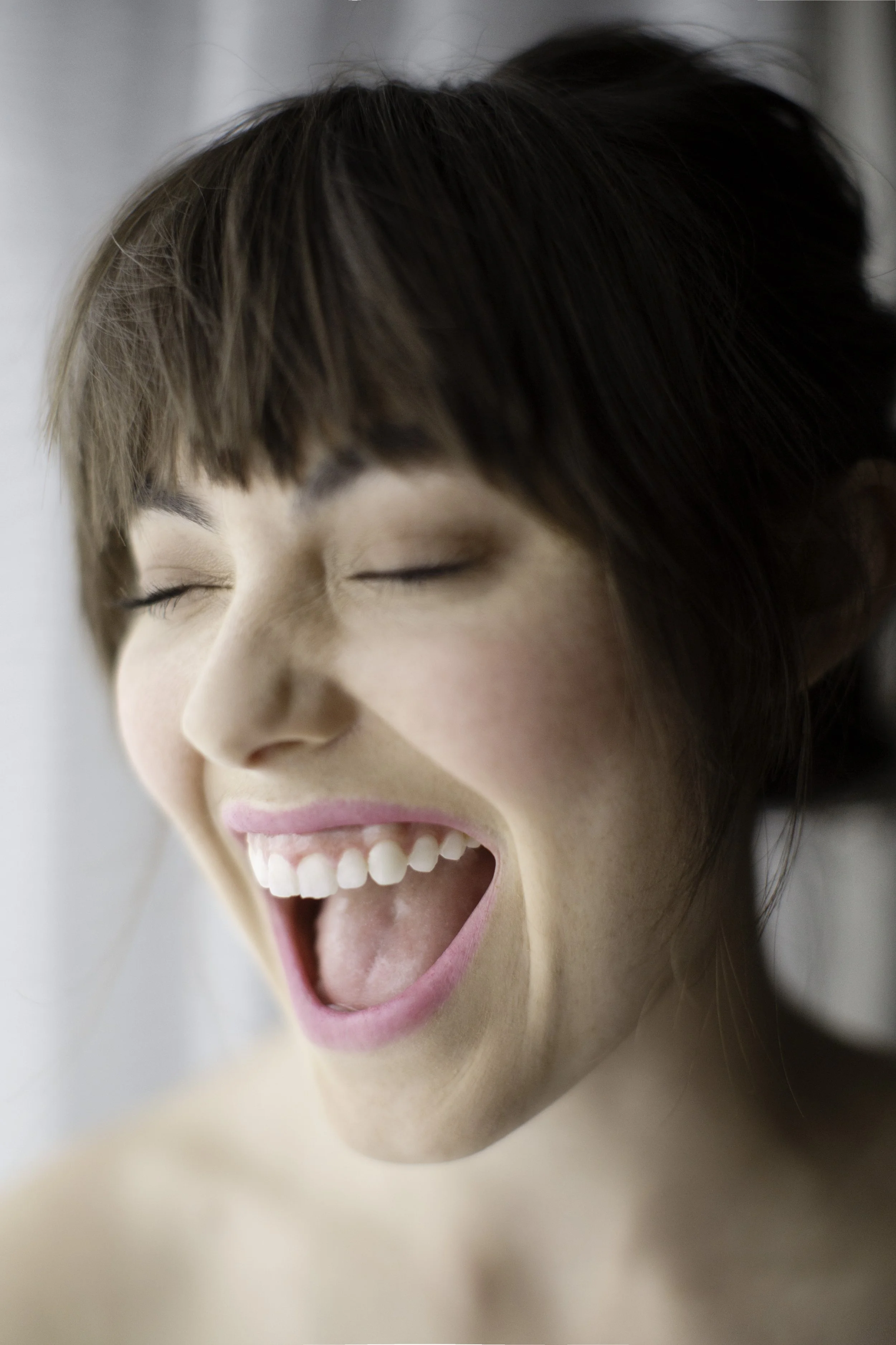 A close-up of a woman laughing or smiling with her mouth open, showing her teeth and tongue, with her eyes closed and light brown hair.