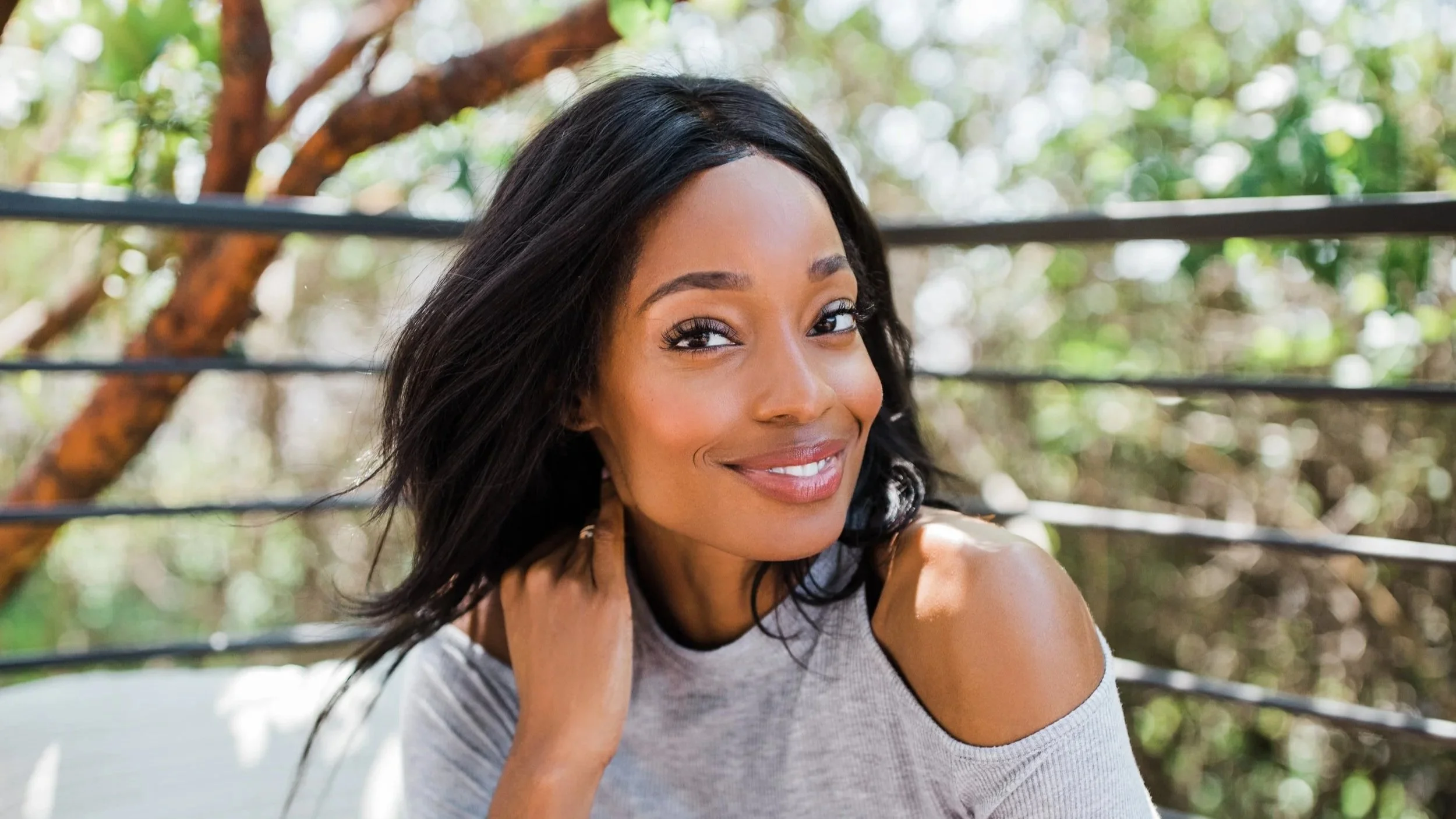A woman with dark hair wearing a light-colored top smiling outdoors with trees and greenery in the background.