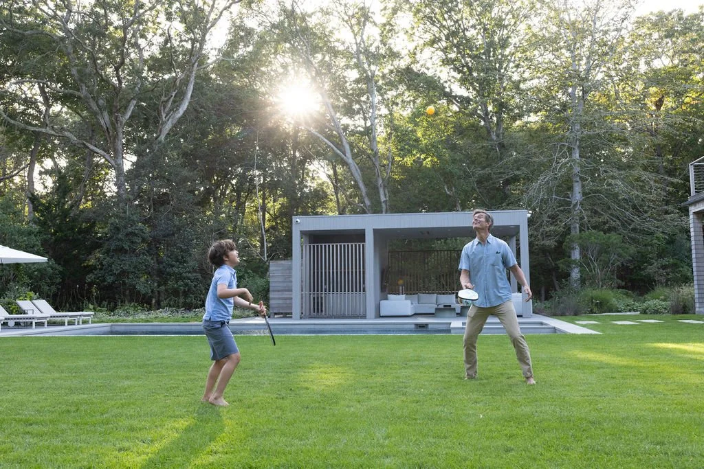 A young boy and man playing golf in a backyard with a modern outdoor seating area, green grass, and trees in the background.