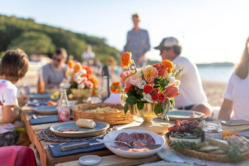 A beachside outdoor brunch table with a floral centerpiece, plates of food, and people enjoying the meal.