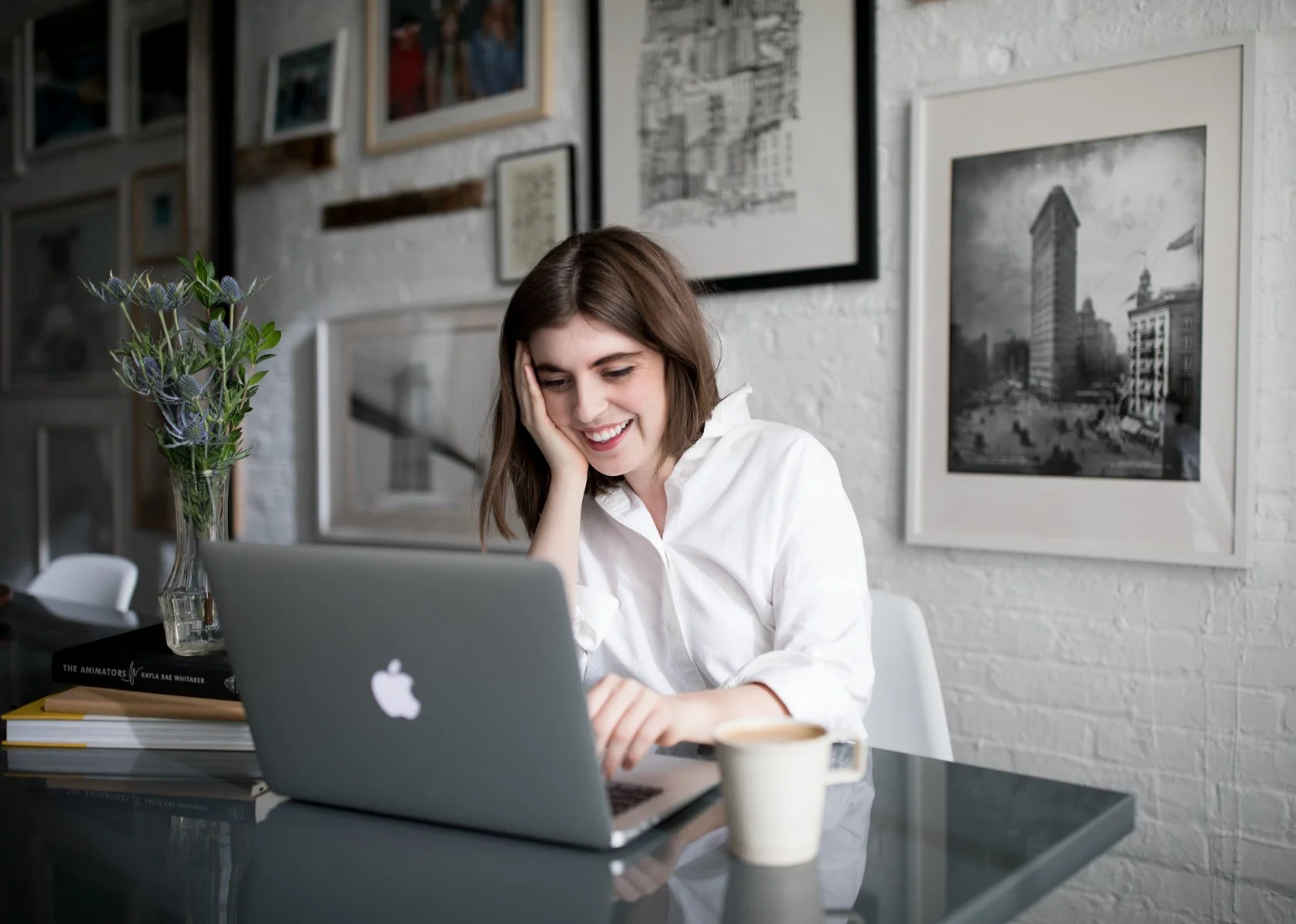A woman sitting at a table working on a laptop in a stylish room with photo art on white brick wall, a flower vase, books, and a coffee cup.