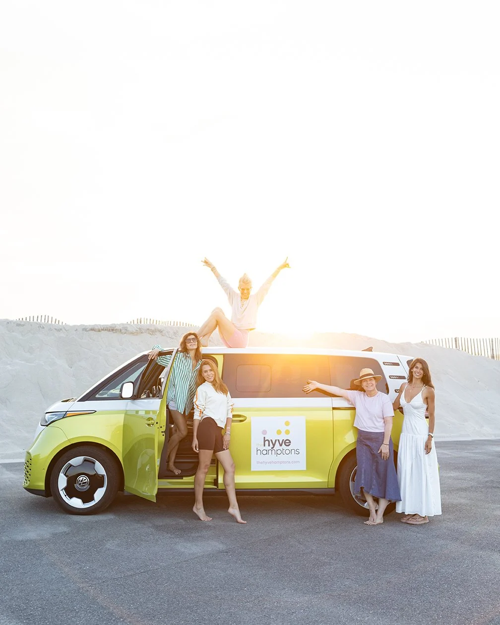 Five women enjoying a sunny day with a yellow and white Hyve Hamptons van at the beach, some sitting on the roof and others standing by the van