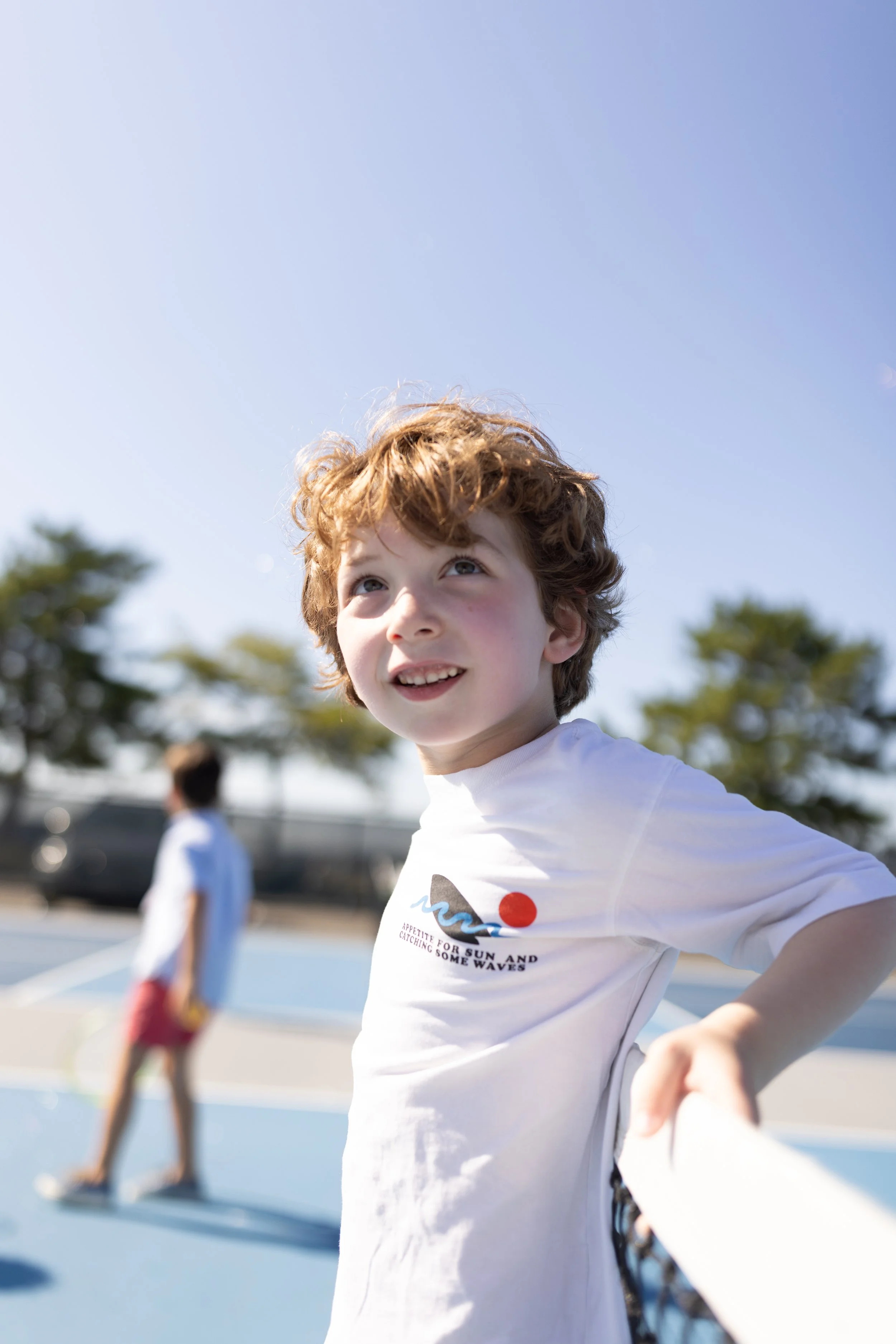 A young boy with curly red hair smiling outdoors at a skate park on a sunny day, wearing a white t-shirt. In the background, other children are skateboarding, and trees can be seen.