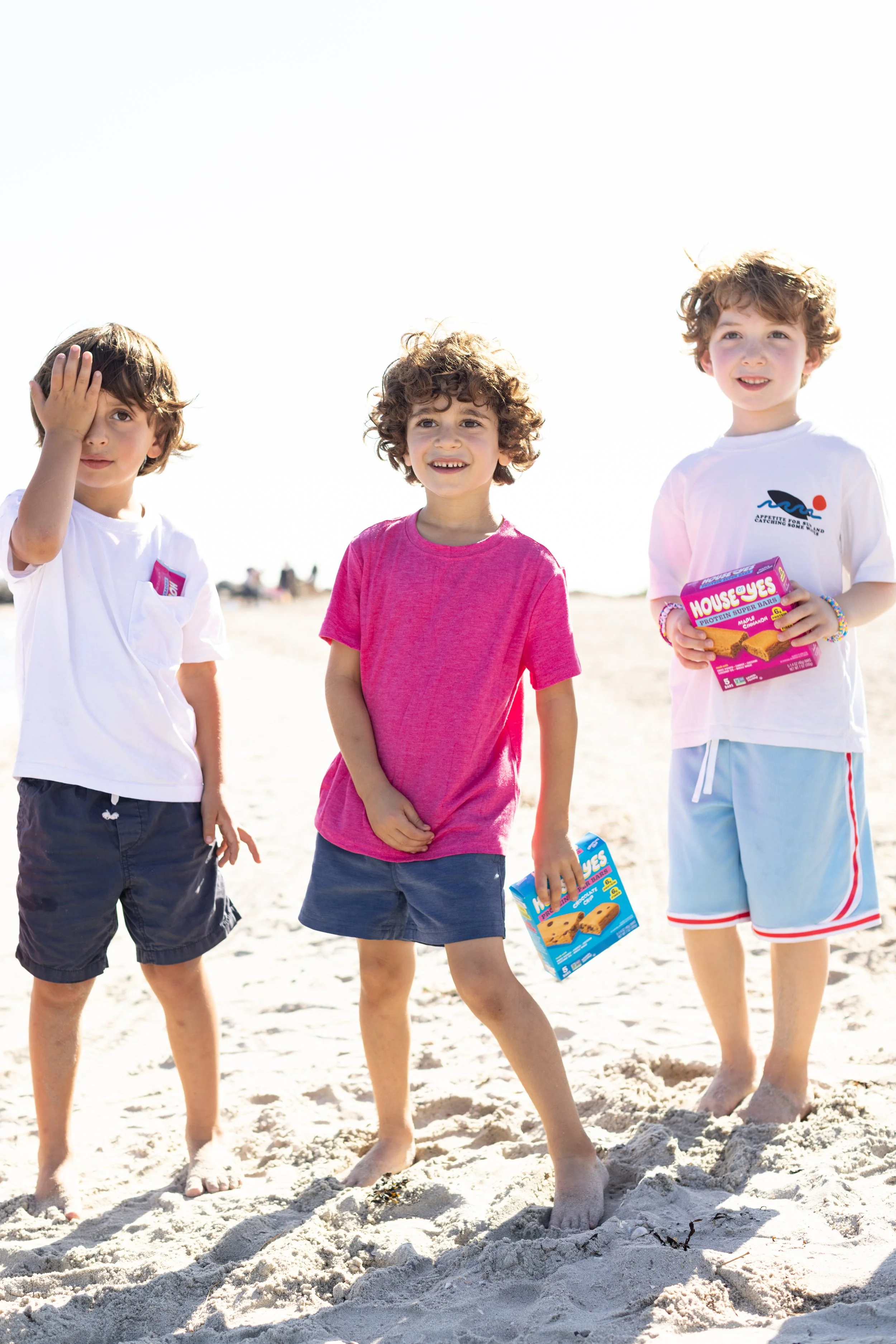 Three young boys standing on the sandy beach, holding boxes of cookie bars, with the sun bright in the sky.