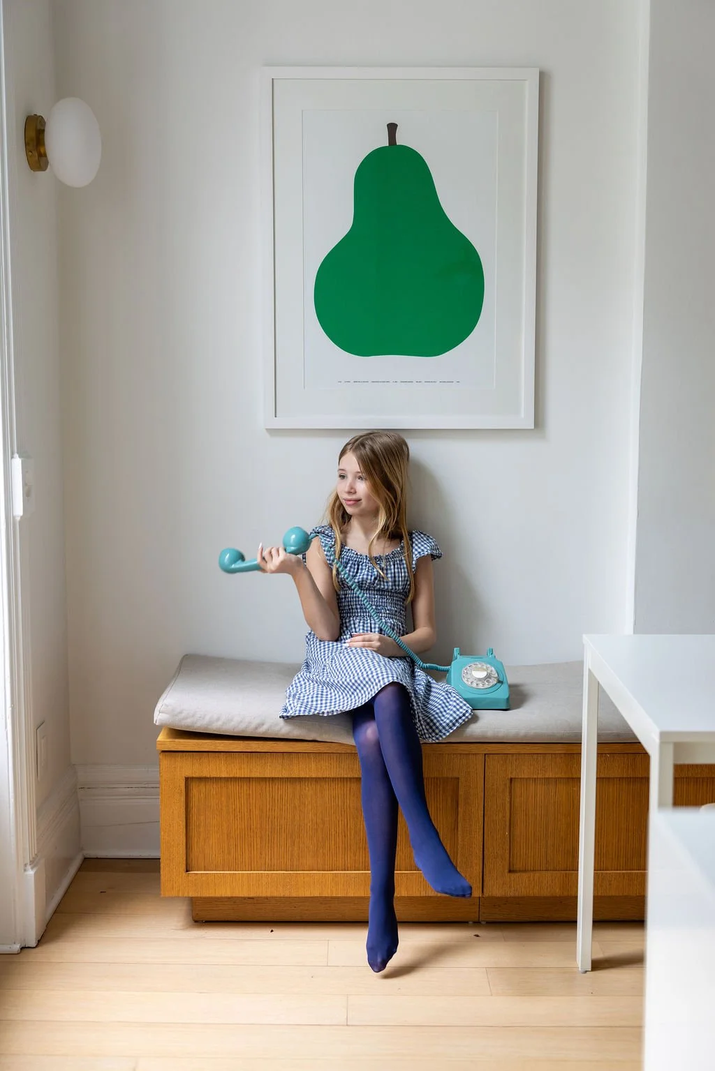 A young girl in a blue checkered dress and blue tights sitting on a wooden bench with a light cushion, holding a vintage teal rotary phone to her ear, in a room with white walls, a framed art print of a green pear above her, and a white side table.