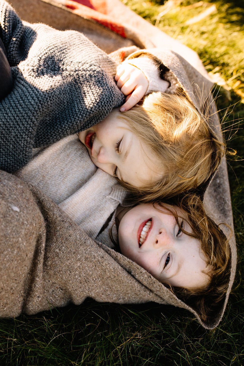 Two children with red hair and freckles laying on the grass under a blanket, smiling and cuddling together during sunset.