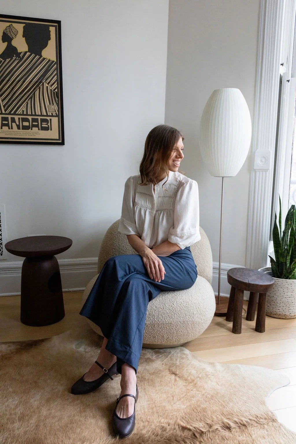 A woman with brown hair smiling, sitting on a beige textured chair, dressed in a white blouse and blue pants, inside a bright room with natural light from the window. There is a tall white lamp, small wooden stools, a potted plant, and a framed art p
