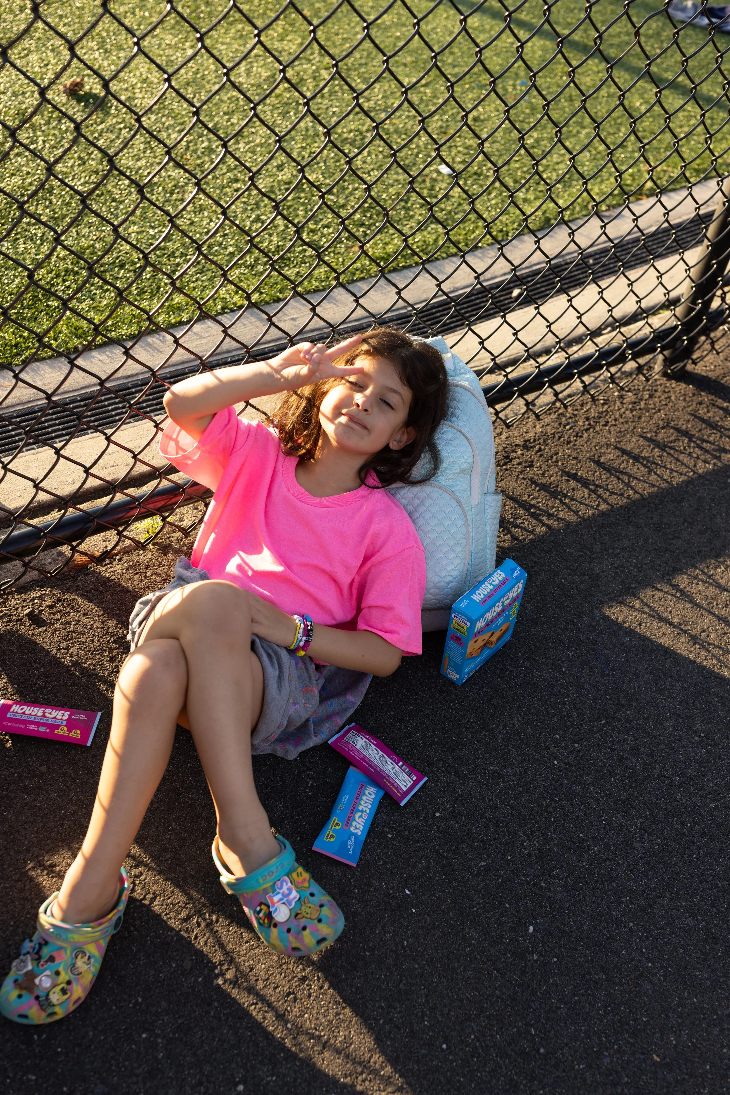 A young girl with brown hair and a pink shirt sitting on a tennis court, making a peace sign with her fingers. She is next to a black chain-link fence, with boxes of 'House Ewe's' snacks nearby, and a backpack behind her. The scene is lit by sunlight