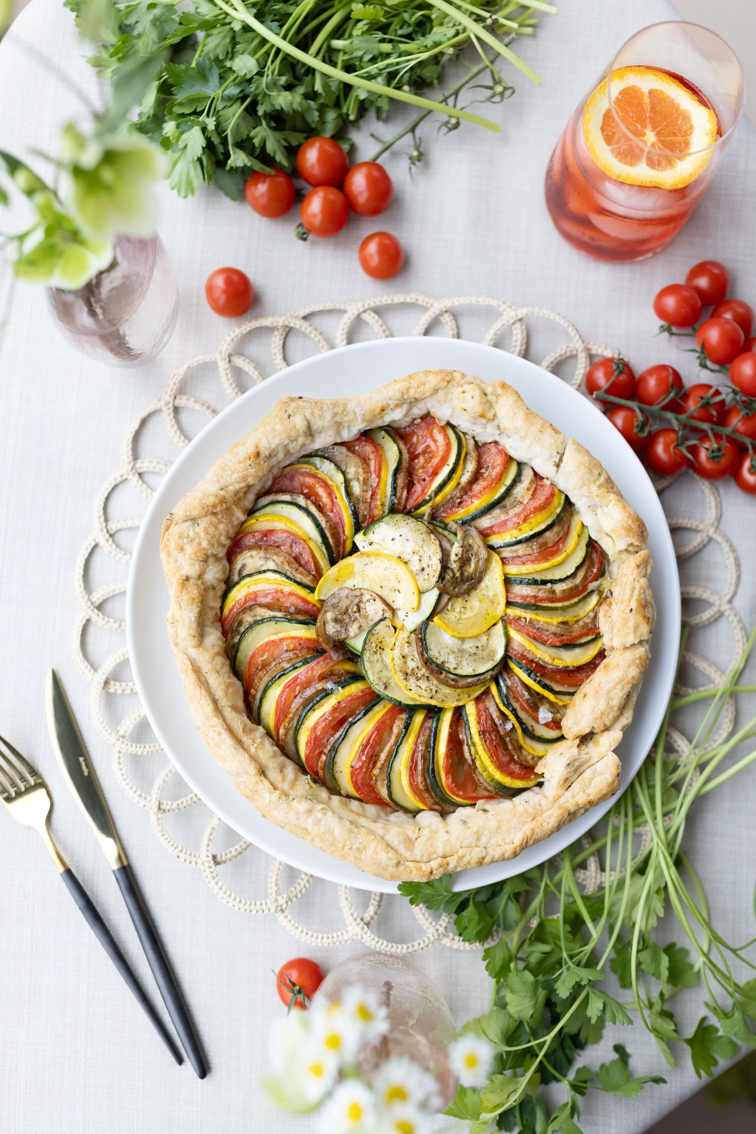A vegetable tart with sliced zucchini, yellow squash, tomatoes, and mushrooms on a white plate surrounded by cherry tomatoes, fresh herbs, and a glass of iced tea with an orange slice, on a light tablecloth.