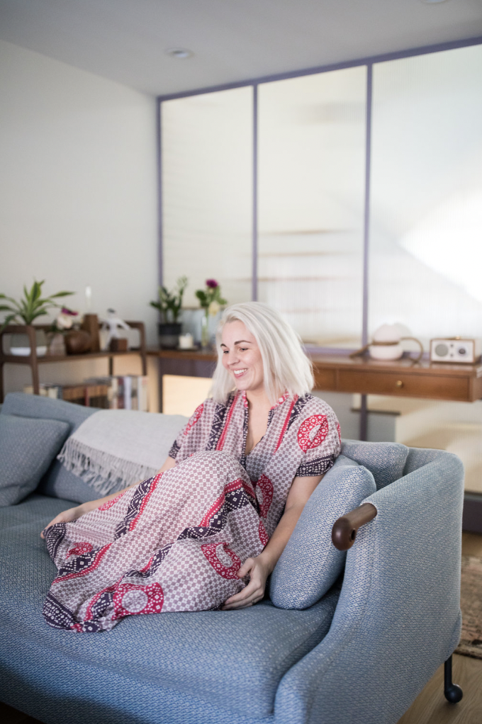 Smiling woman with bleached blonde hair sitting on a blue sofa in a bright living room, wrapped in a patterned blanket.