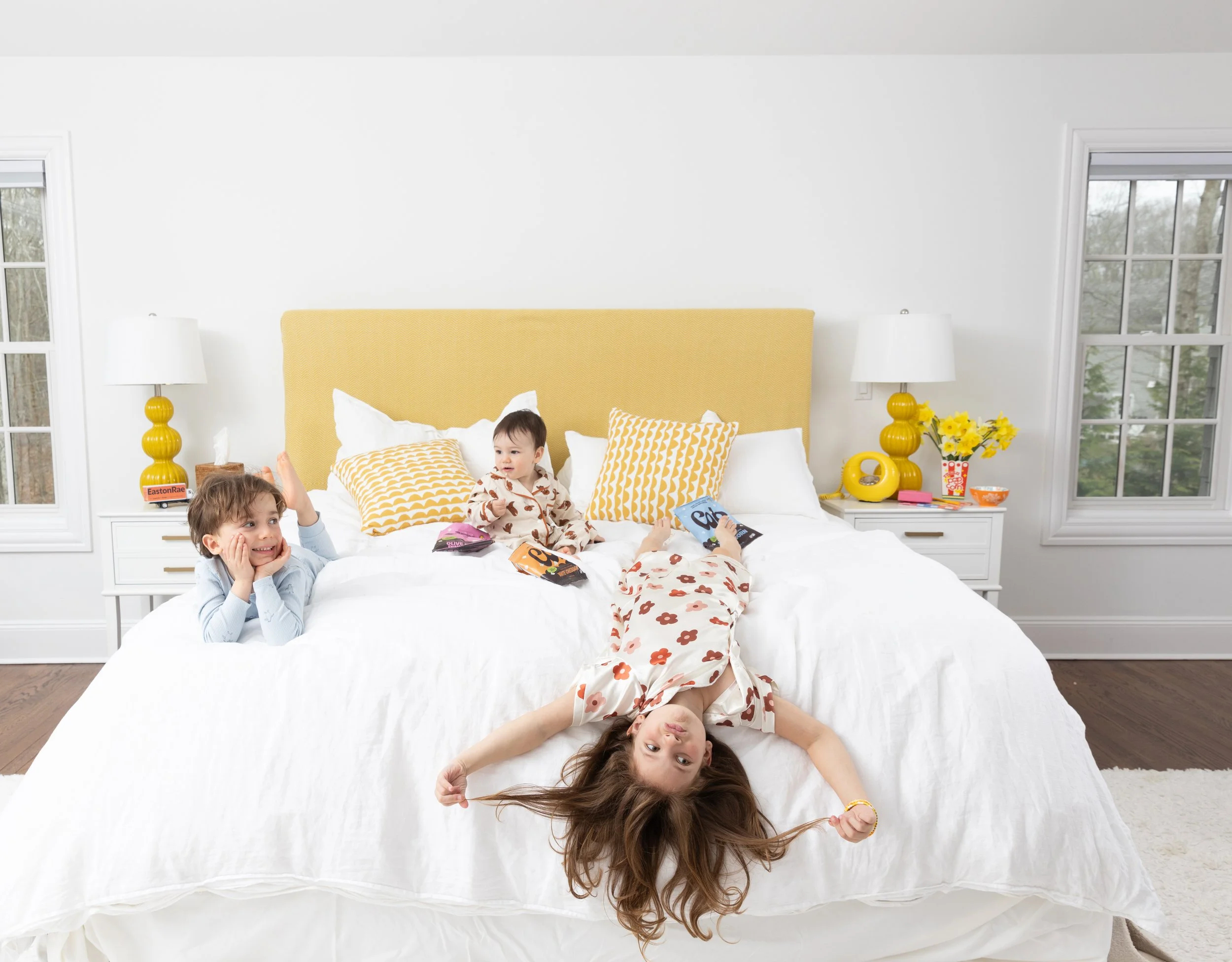 Multiple children are on a large white bed in a bright bedroom with white walls and large windows. One girl with long hair is lying on the bed, while two children are sitting and playing among pillows.