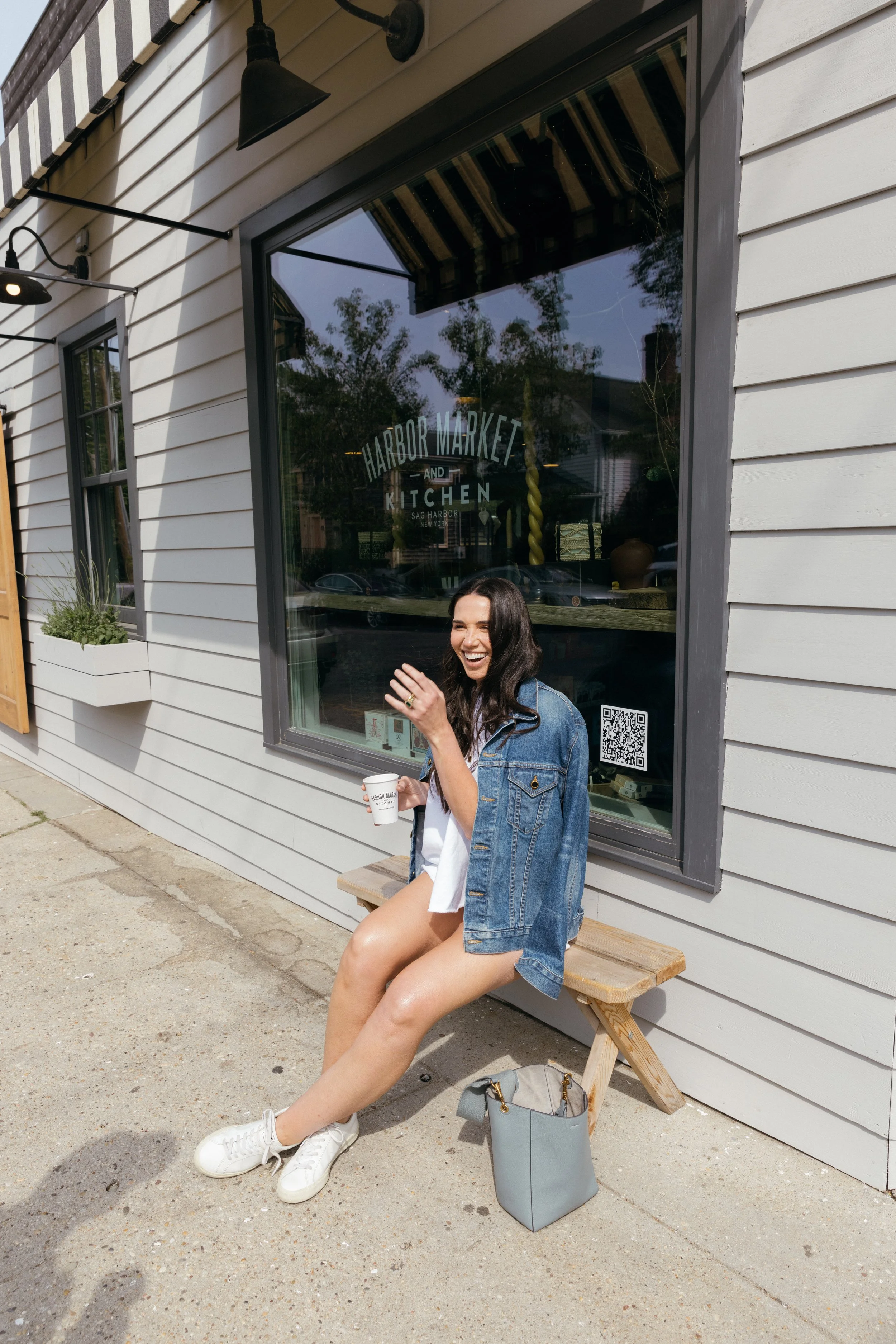 A woman sitting on a wooden bench outside of a storefront, smiling and laughing while holding a cup. She is wearing a denim jacket, white dress, and white sneakers. A gray handbag is on the ground beside her.