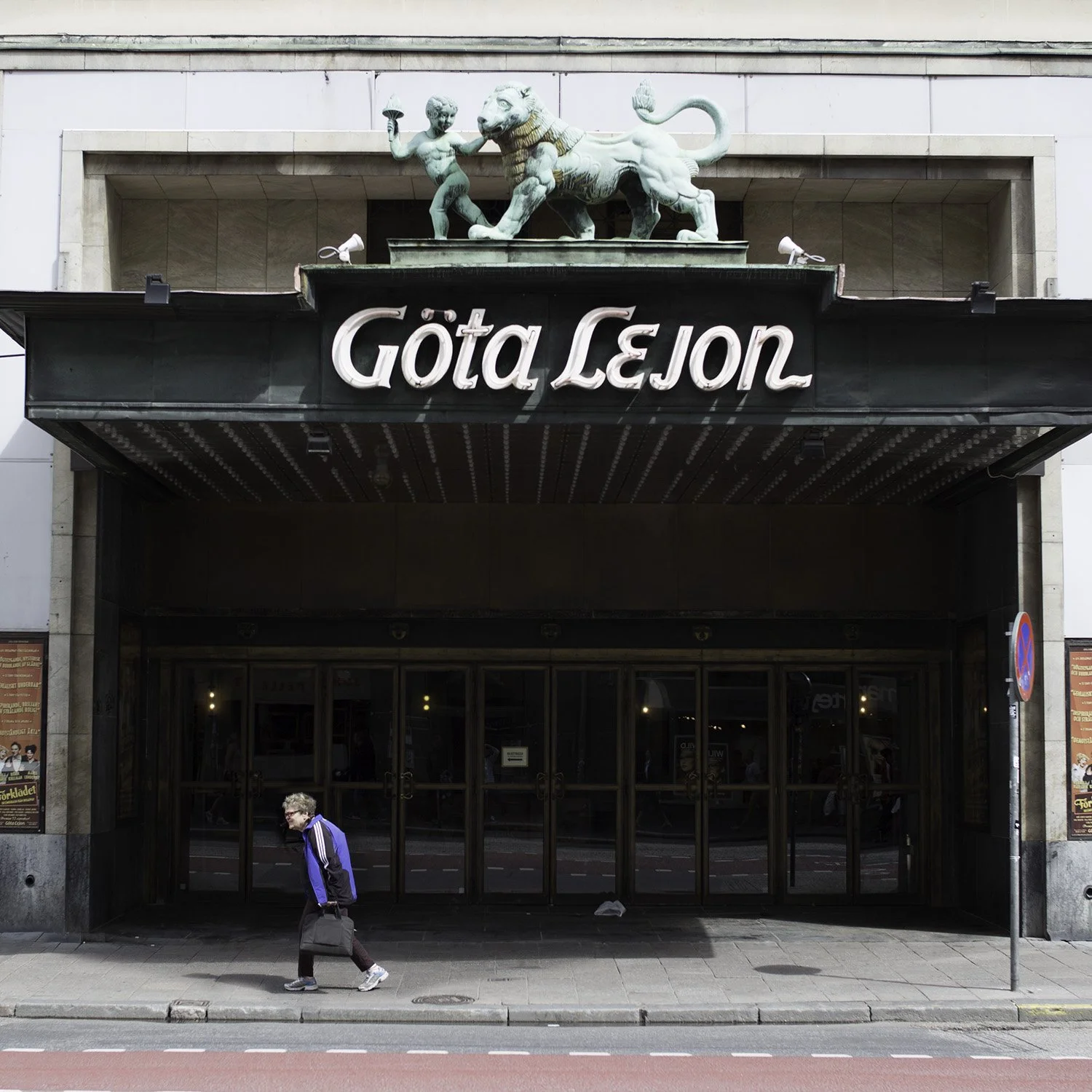 Street view of a theater entrance labeled 'Gøta Lejon' with a lion sculpture and a child walking by in front.