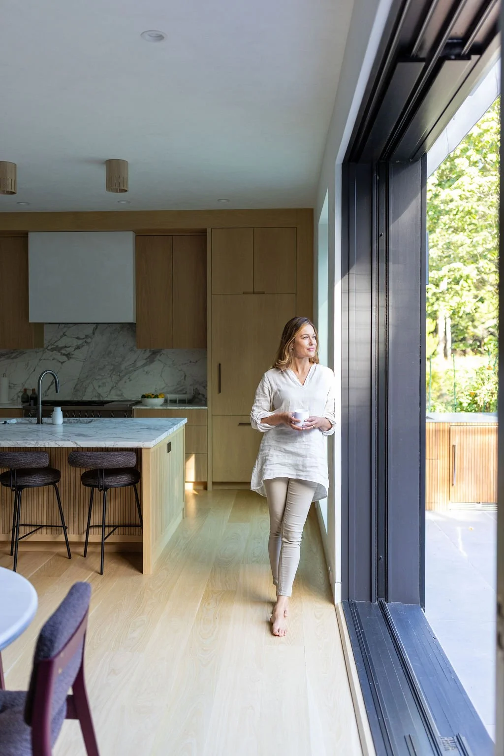Woman standing barefoot by large sliding glass door in modern kitchen, holding a mug, looking outdoors during daytime.