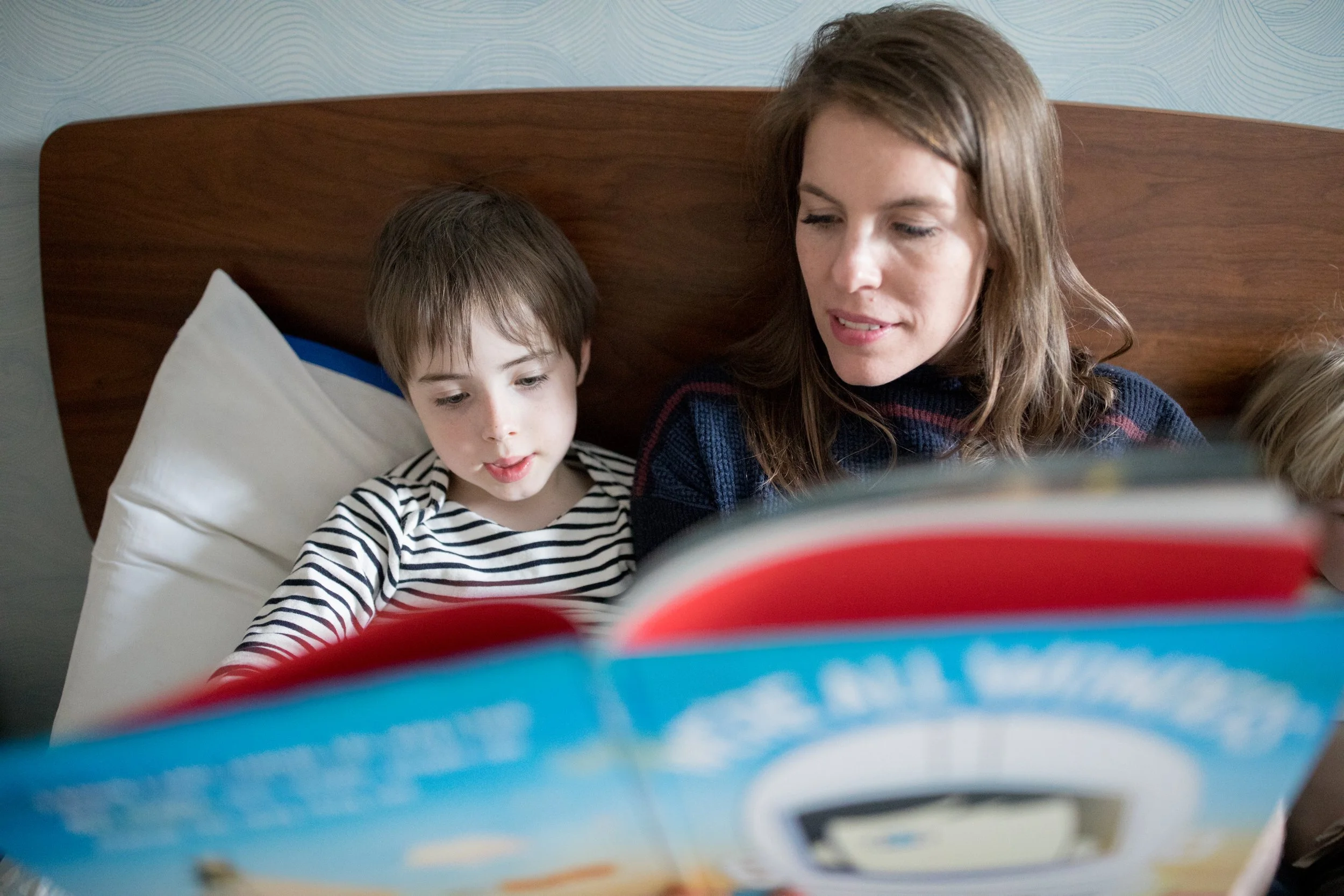 A woman and a young boy sitting together on a bed, looking at a colorful book.