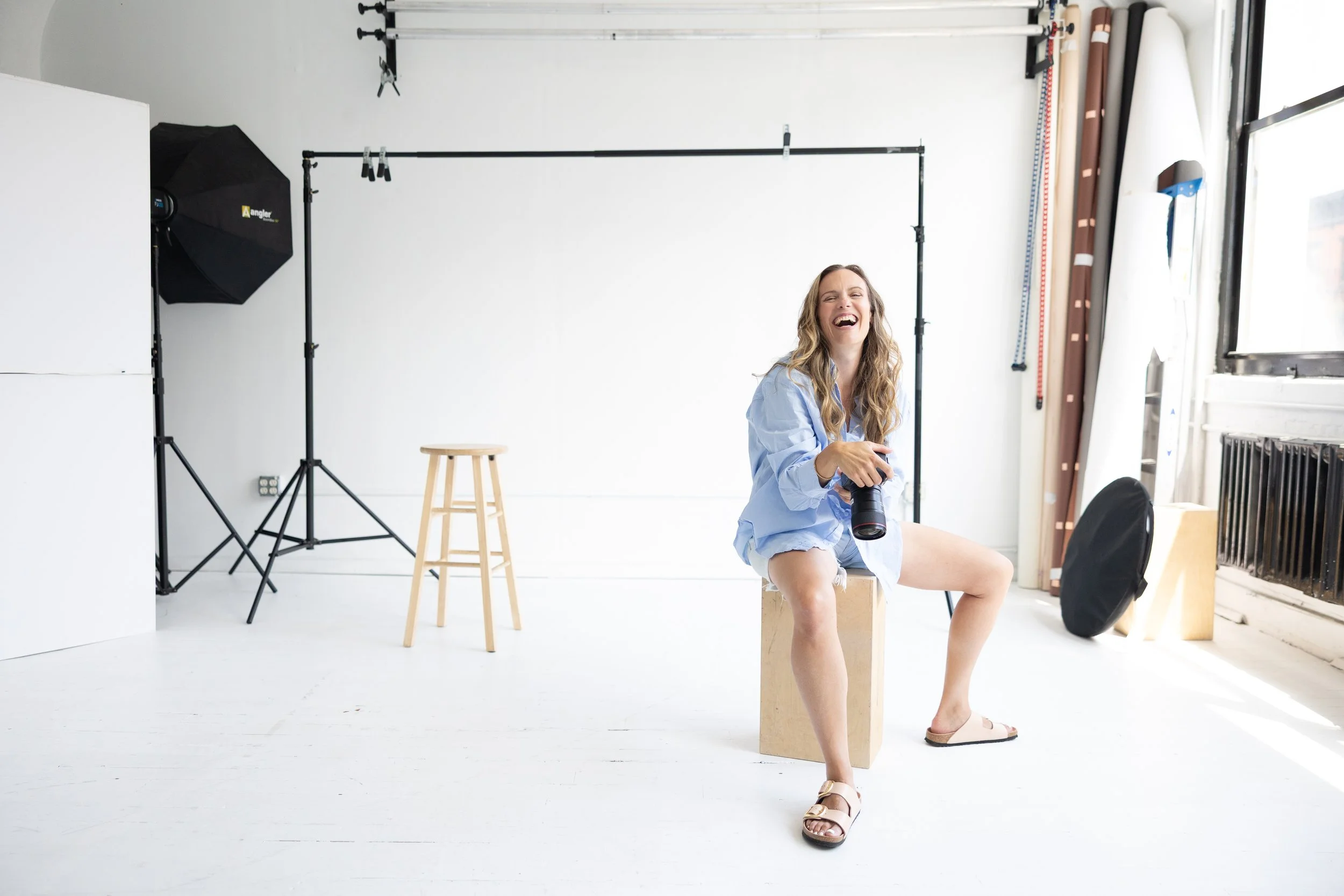 A woman sitting on a wooden box in a photography studio, laughing and holding a camera, surrounded by photography equipment, with a white backdrop and natural light coming through large windows.