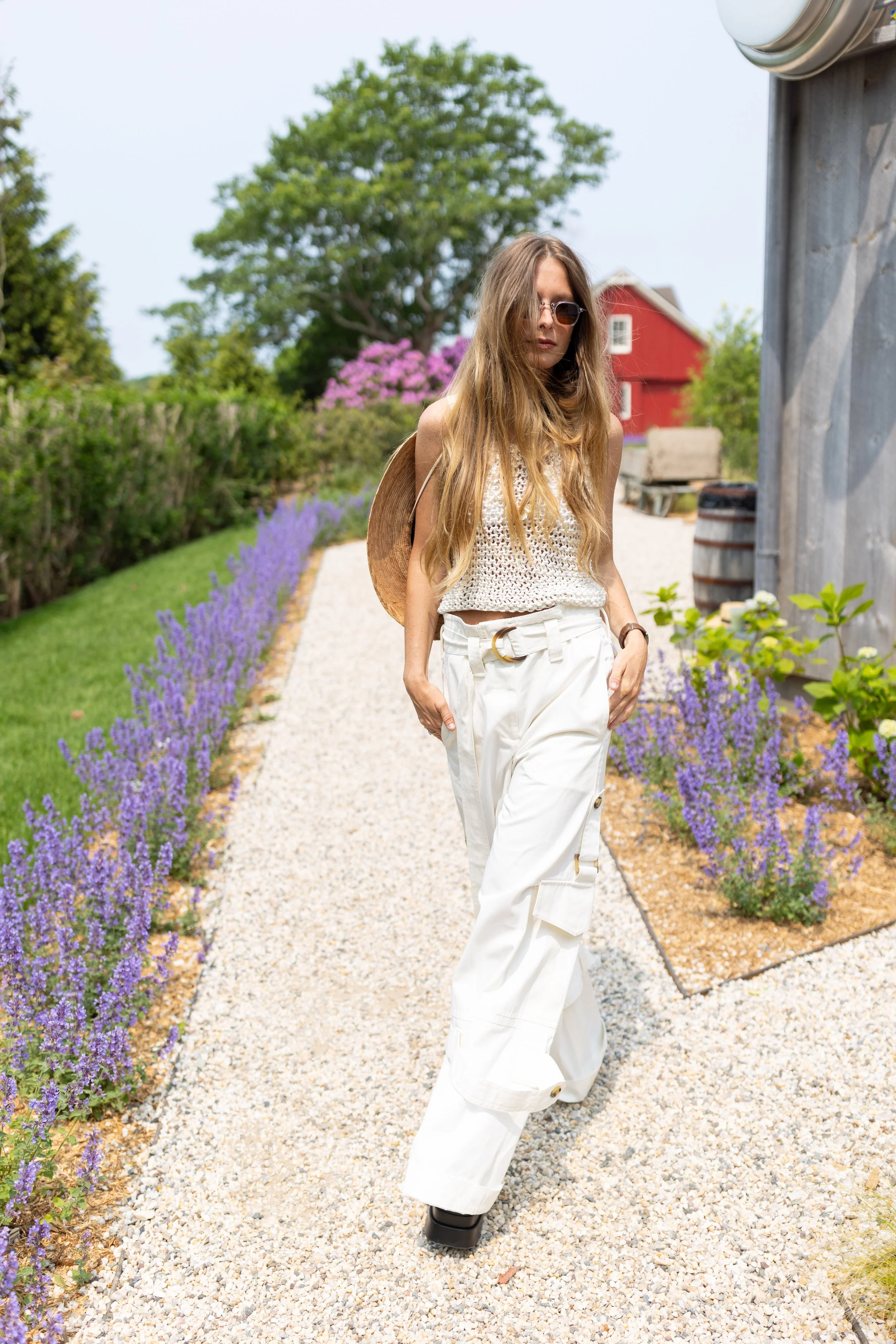 Young woman with long hair wearing sunglasses, a beige top, and white cargo pants, walking on a gravel garden path lined with purple flowers, with trees and a red barn in the background.