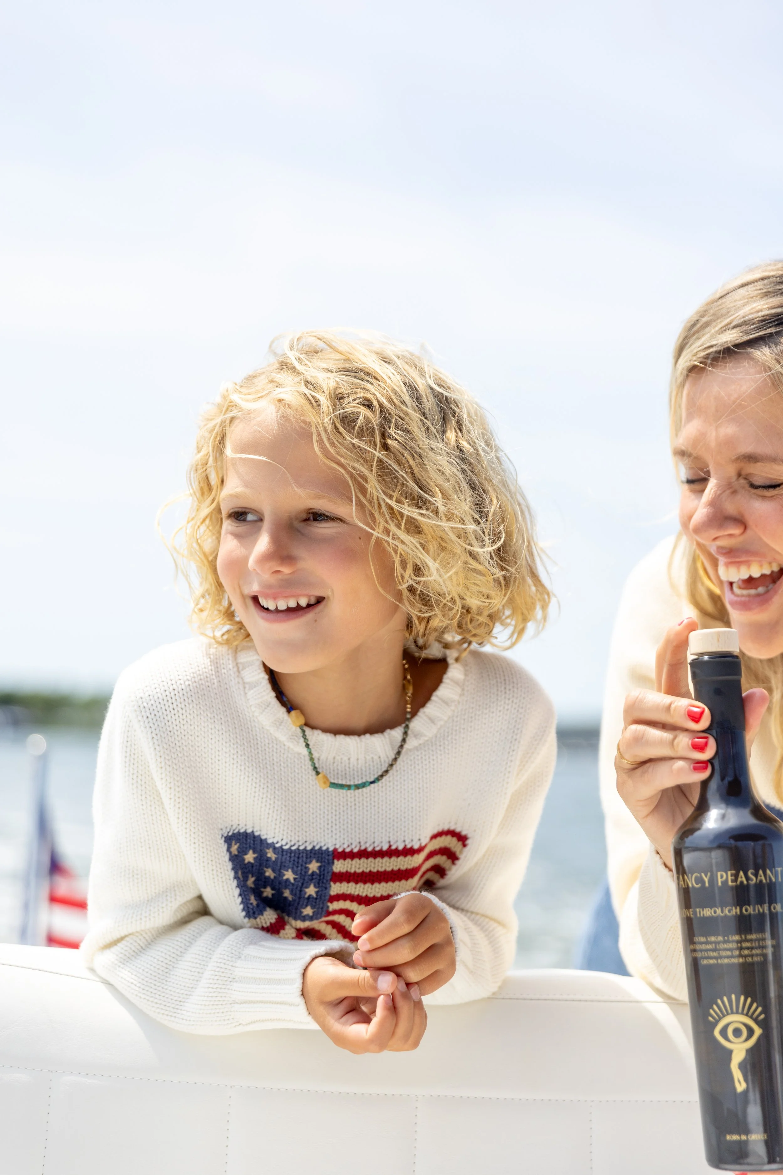 A young girl with curly blonde hair wearing a sweater with an American flag on it, smiling and looking to her left. An adult woman with blonde hair and red nail polish also smiling, holding a bottle of olive oil. The background shows an outdoor setti