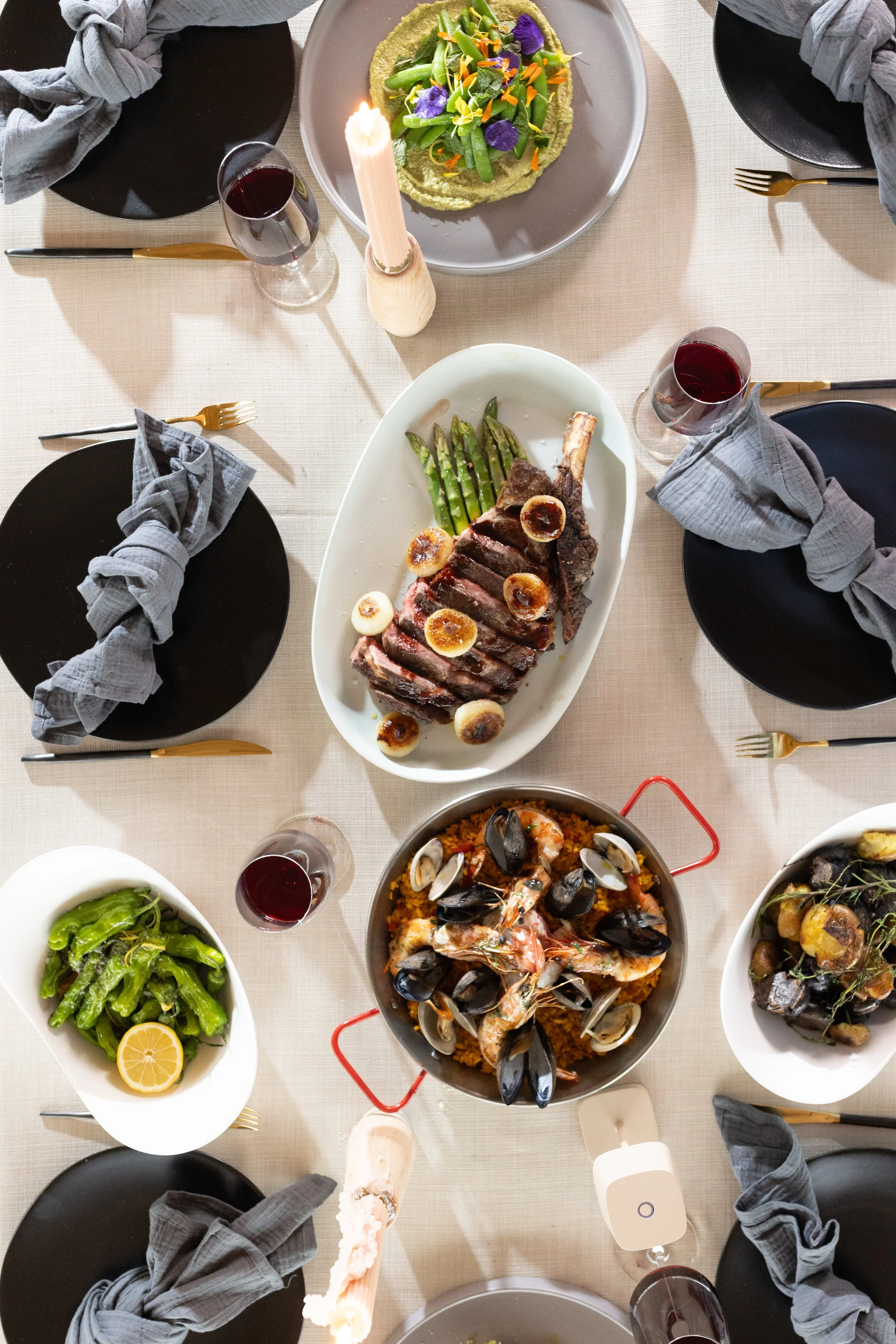 Overhead view of a dining table set with black plates, gold utensils, grey cloth napkins, glasses of red wine, and several dishes including a large grilled meat with garlic, grilled asparagus, a seafood paella with shrimp, mussels, and clams, a green bean dish with lemon, and a vegetable dish topped with herbs.
