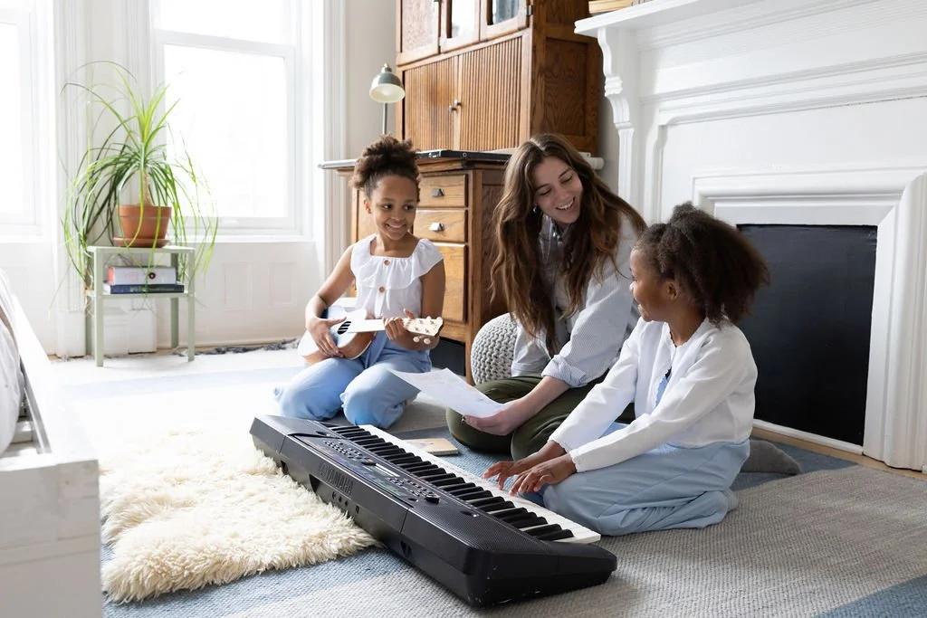 Three people, a woman and two girls, sitting on a rug in a bright living room with large windows. They are engaged in a music activity with a digital keyboard and sheets of paper, one girl playing the keyboard, another girl holding a paper, and the woman smiling at them.