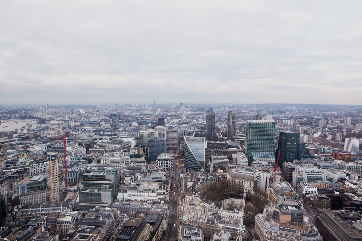 Aerial view of a city with numerous tall skyscrapers, busy streets, and a river in the distance under a cloudy sky.