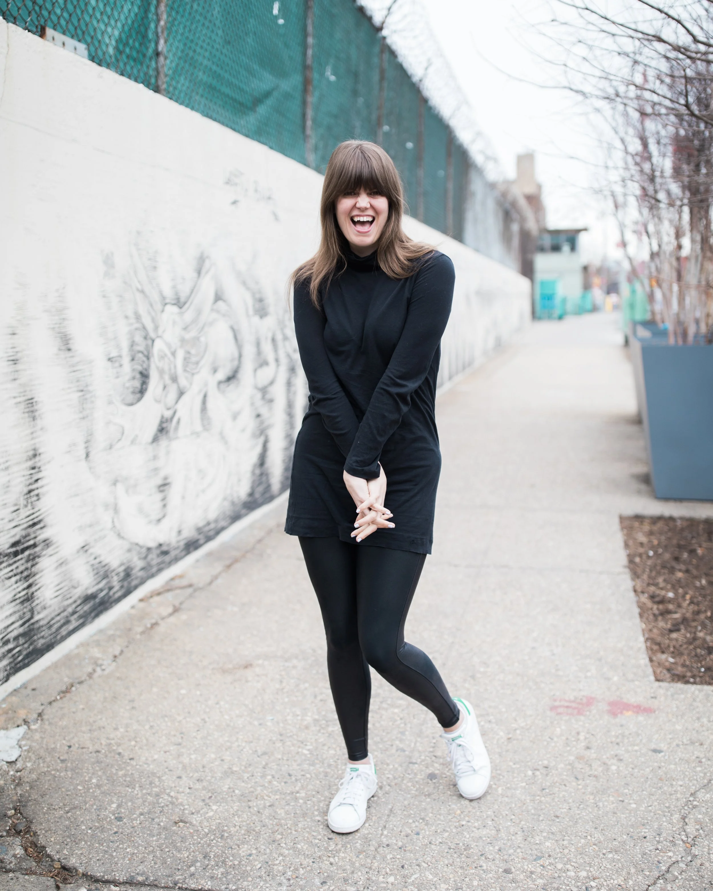 Young woman with brown hair, wearing a black long-sleeve dress, black leggings, and white sneakers, smiling and crossing her arms while standing on a sidewalk next to a painted wall with graffiti, with leafless trees in the background.