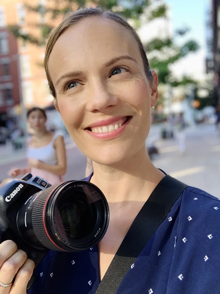 A woman smiling and holding a Canon camera, outdoors in an urban area with buildings and trees in the background.