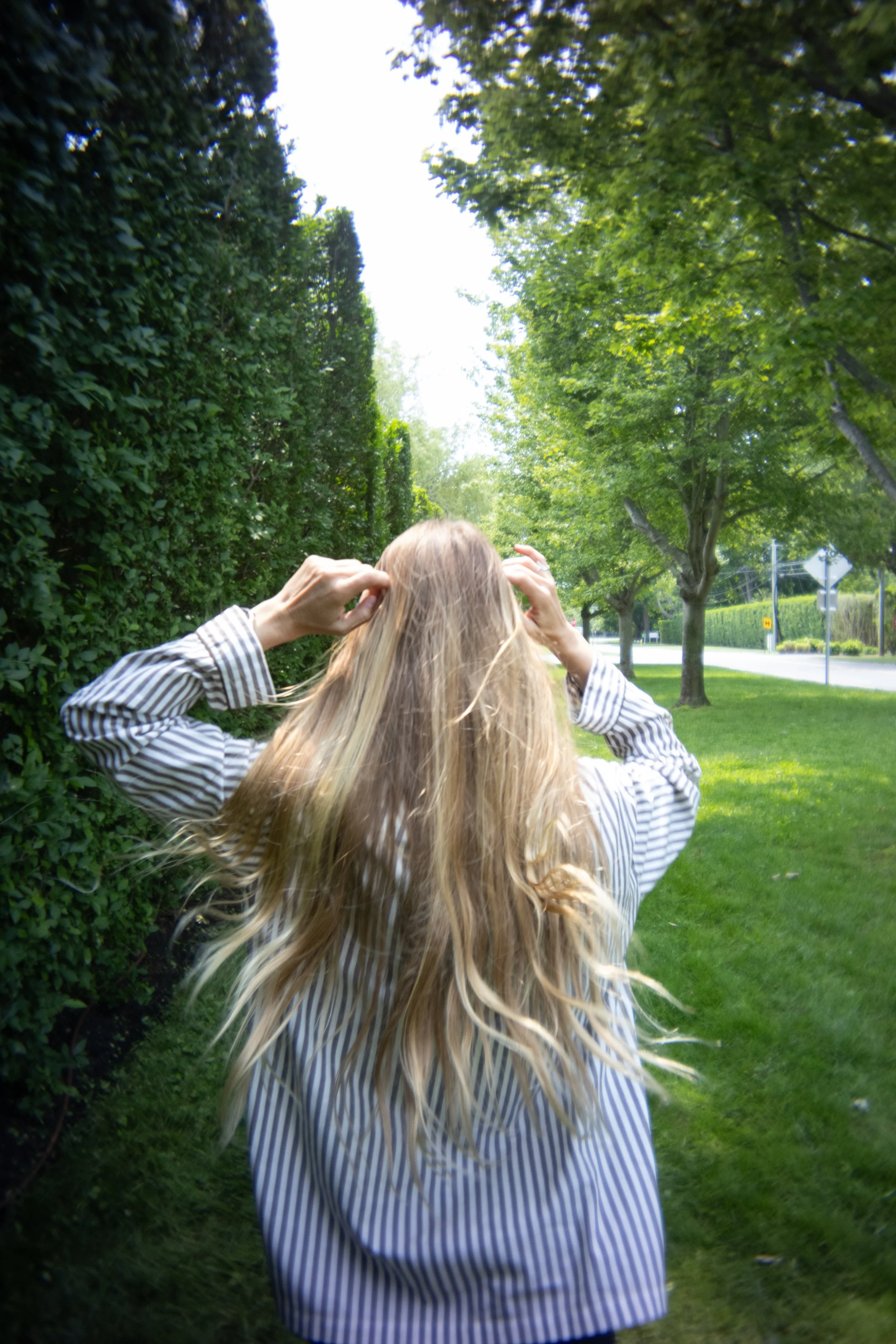 A person with long blonde hair, wearing a striped shirt, standing on a sidewalk in a green park or neighborhood with trees and grass, touching their hair.