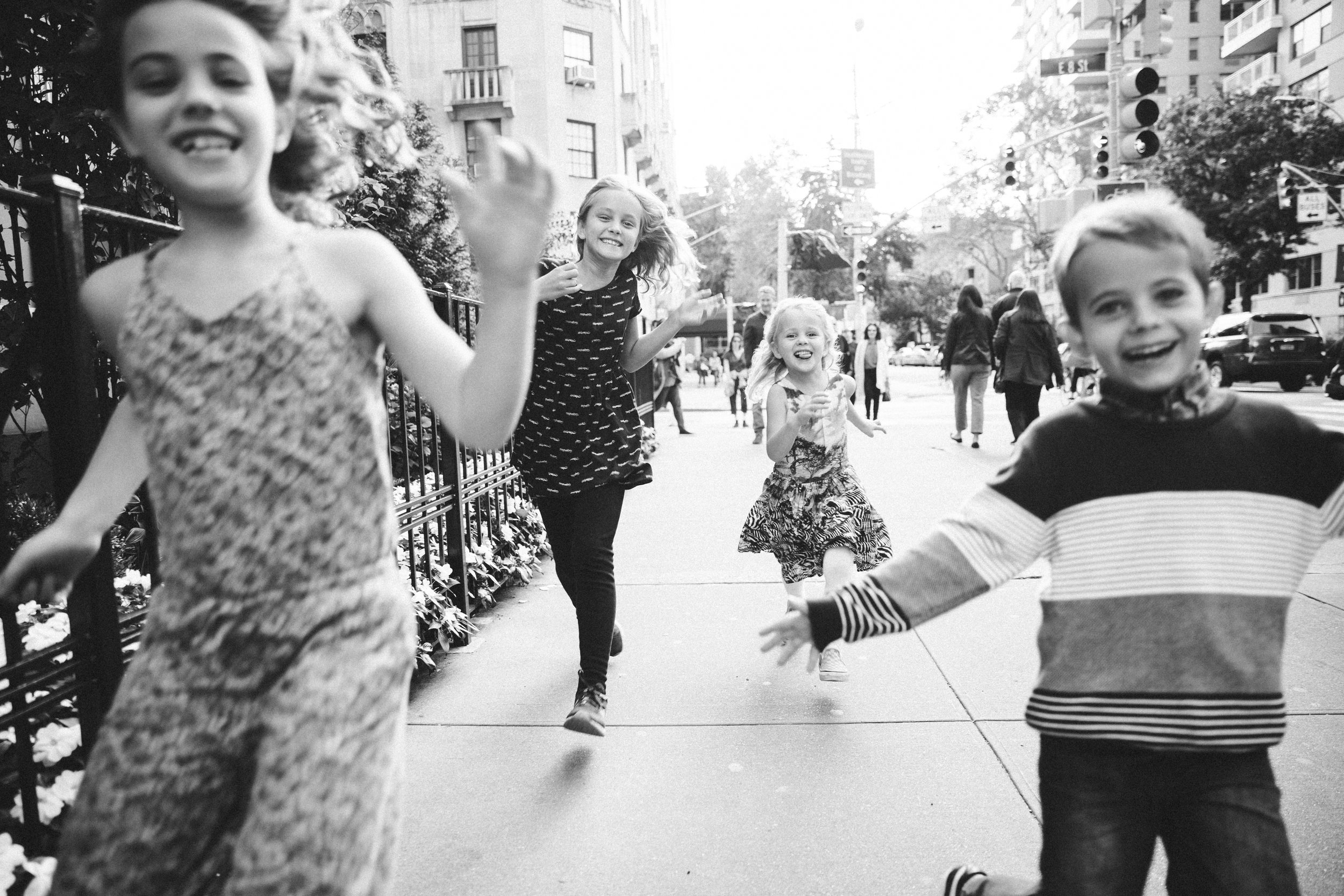 Children running and playing on a city sidewalk. Two girls and a boy in the foreground, with others in the background. The scene is in black and white.