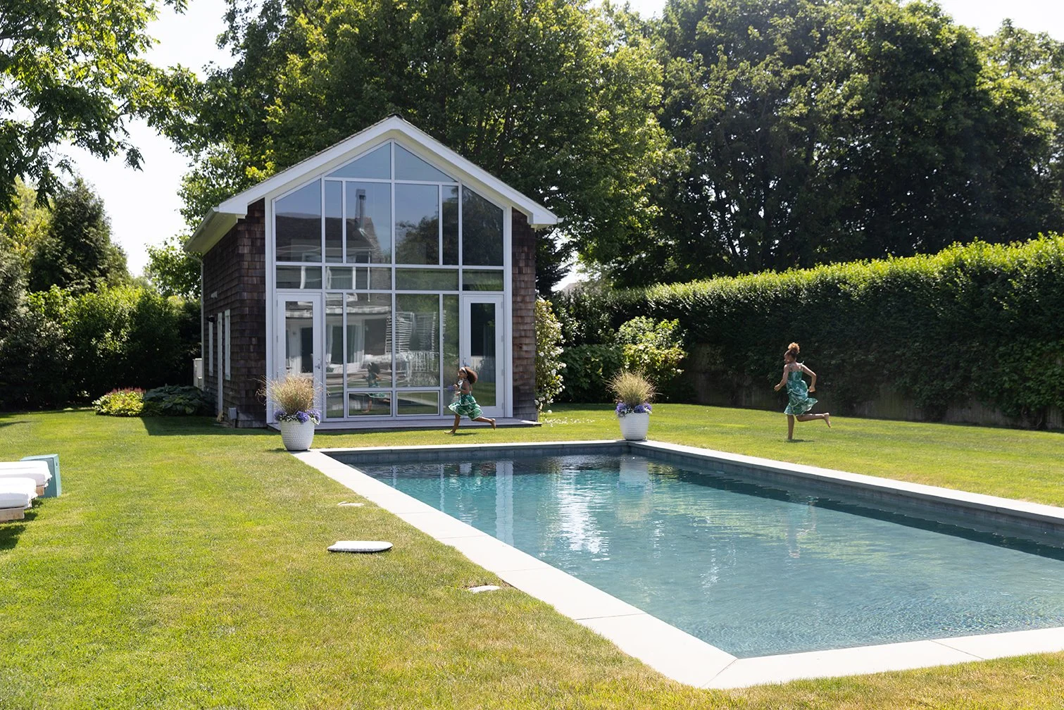 A backyard with a rectangular swimming pool, a grassy lawn, and a modern house with large glass windows. Two girls in summer dresses are running and playing near the pool, and there are potted plants with flowers on the lawn.